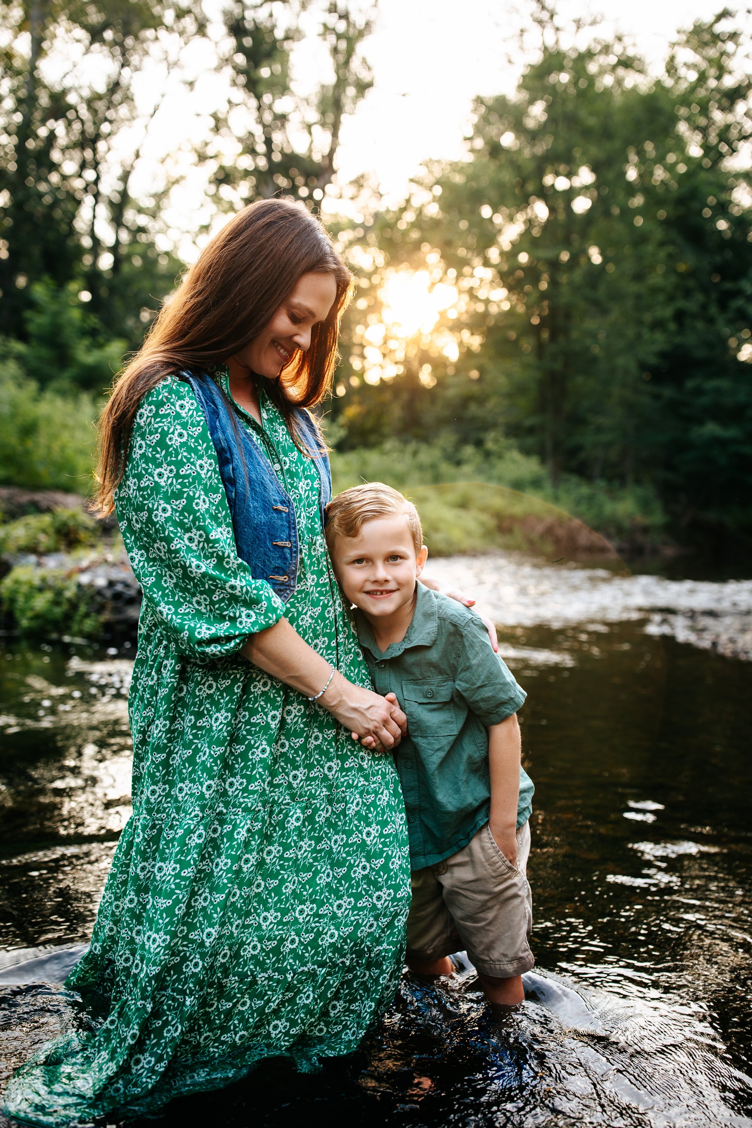 Mom in green dress looking at her son while he is looking at the photographer's camera during their river session
