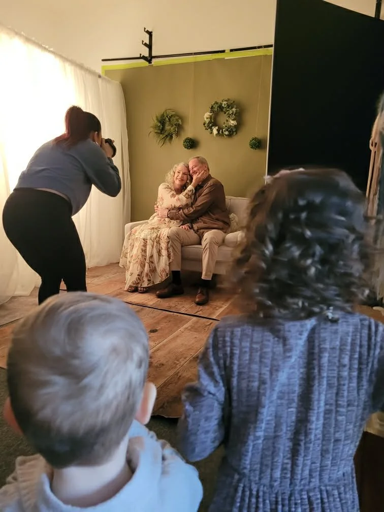 A photo of an elderly couple seated on a white couch, celebrating a special moment while a photographer takes their picture. Two children with light-colored hair are watching from the foreground, and a woman with curly hair is partially visible on the right. The backdrop features a green wall with three wreath decorations.