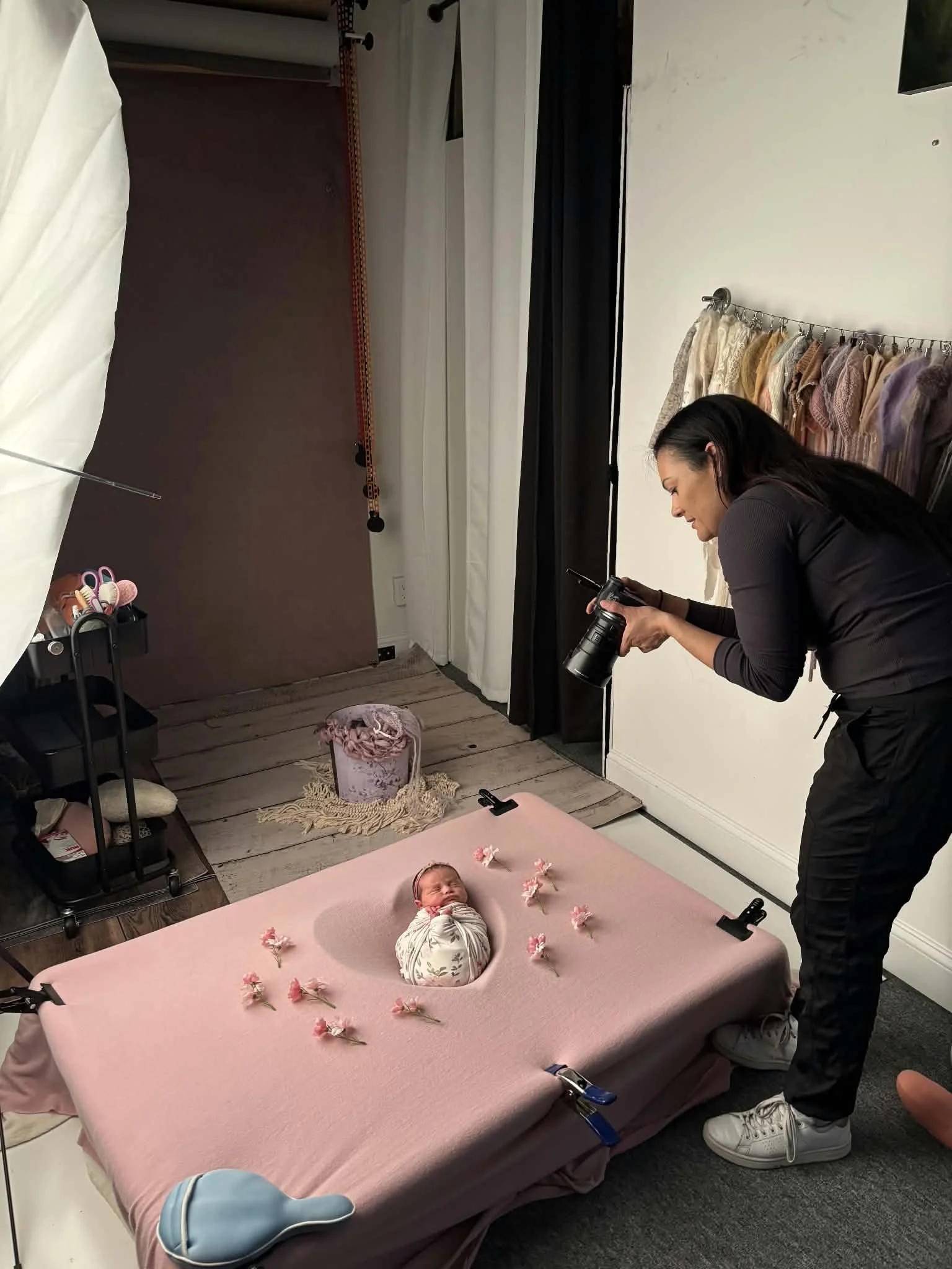 A woman taking a photograph of a newborn baby lying on a pink blanket with pink flowers arranged around, in a photography studio.