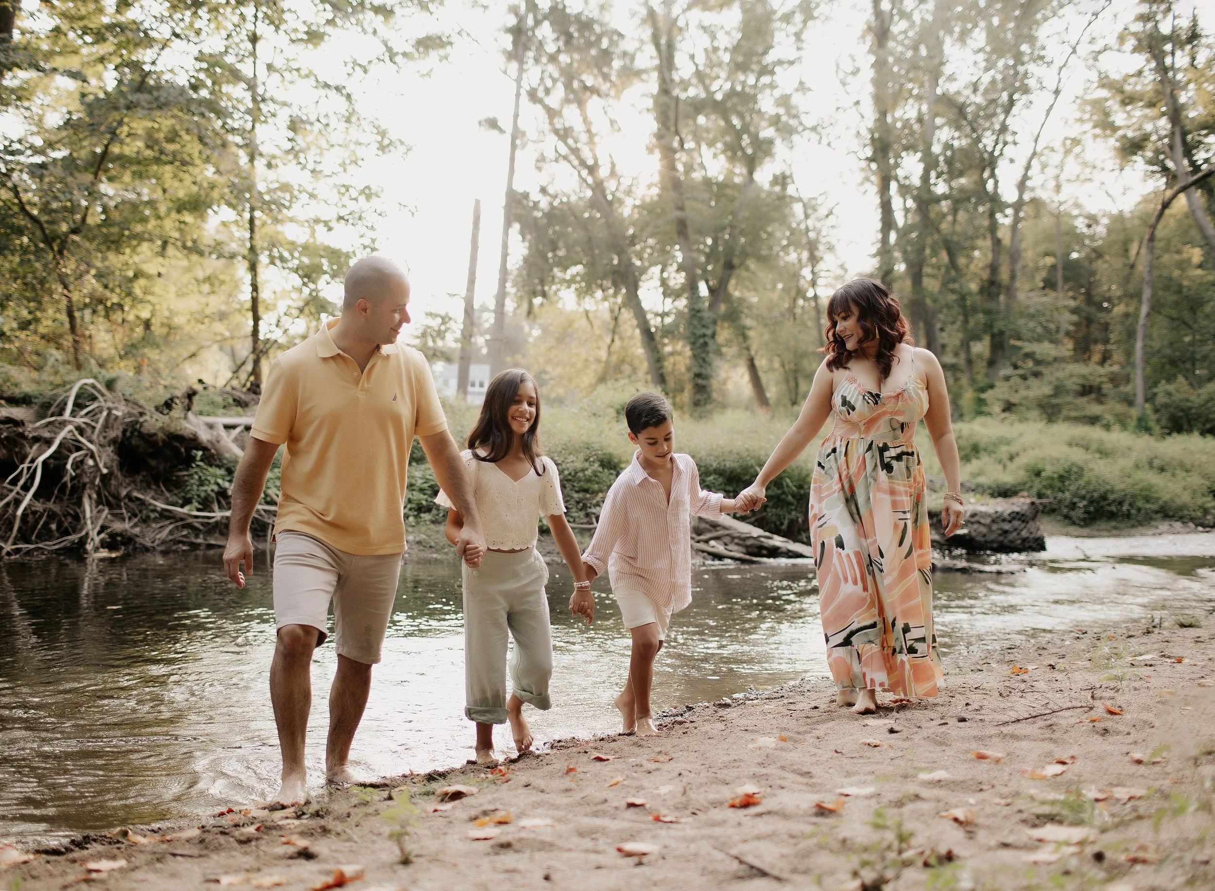 A family of five, including two children, walks hand-in-hand through a shallow river in a wooded area during late afternoon or early evening, with sunlight filtering through the trees.