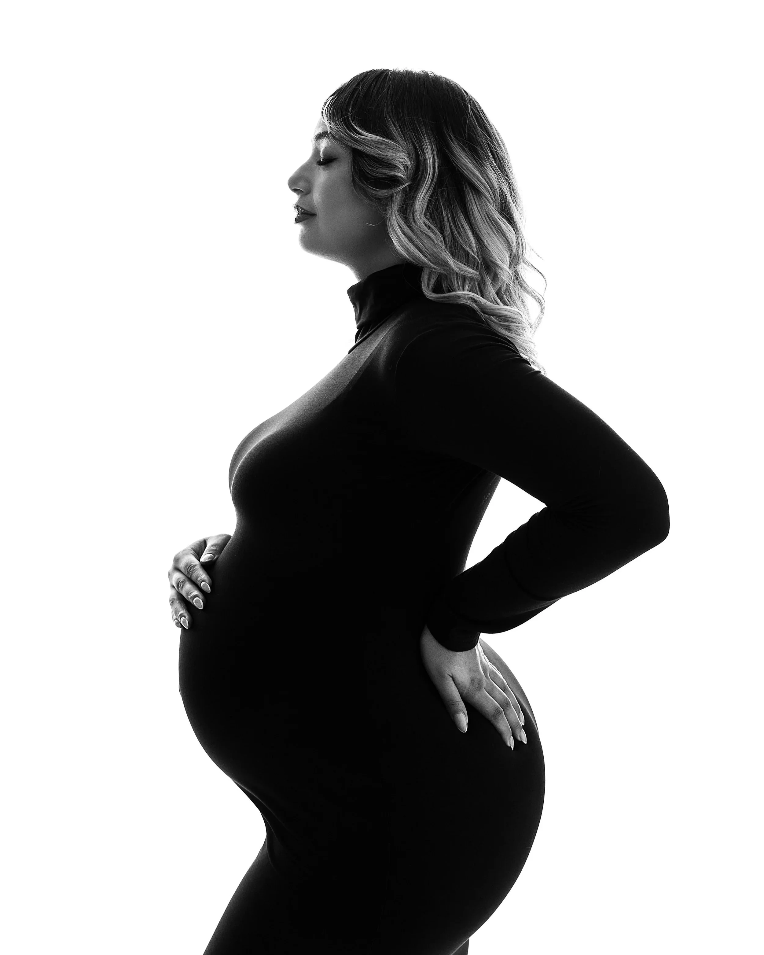 Profile of pregnant woman with a black dress on a white background touching her belly with eyes closed in Lincoln Park, NJ photography studio
