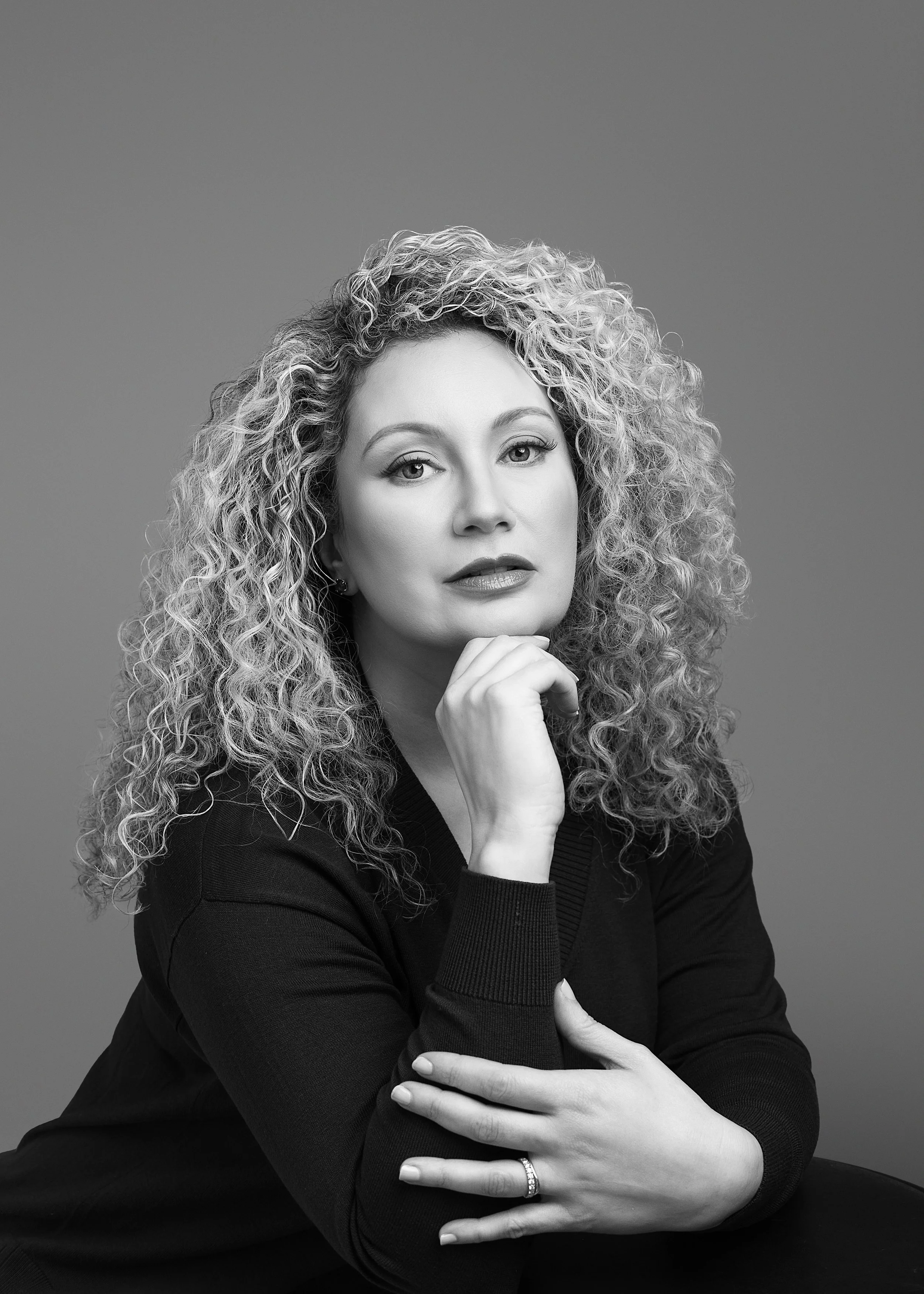 Balck and white beauty headshot of woman with blonde curly hair posing her head on her hand looking at the camera