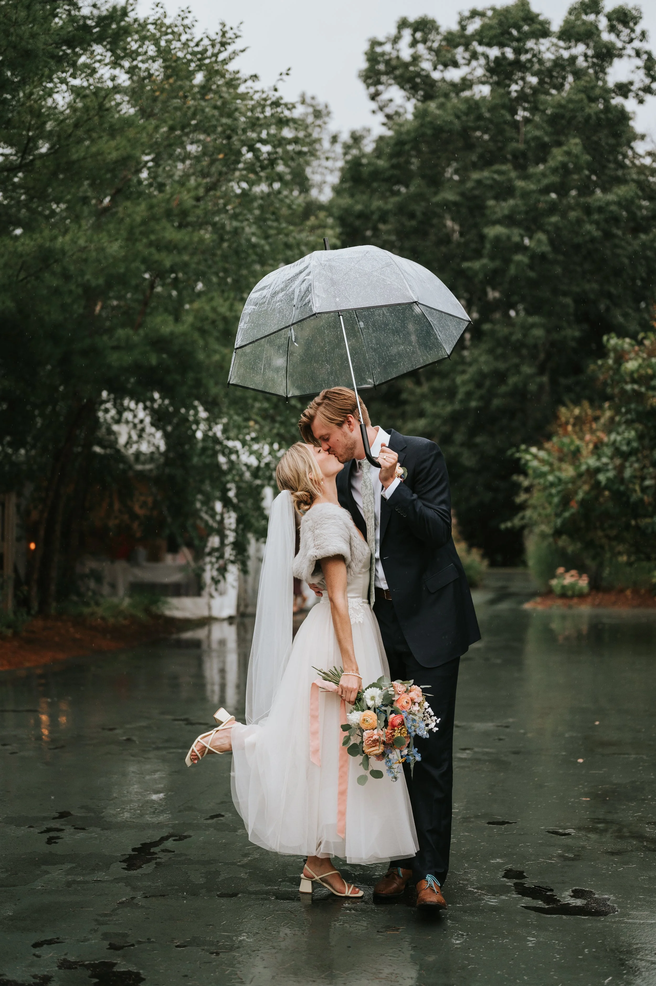 A couple dressed in wedding attire sharing a kiss under an umbrella on a rainy day, standing on wet pavement with trees in the background.