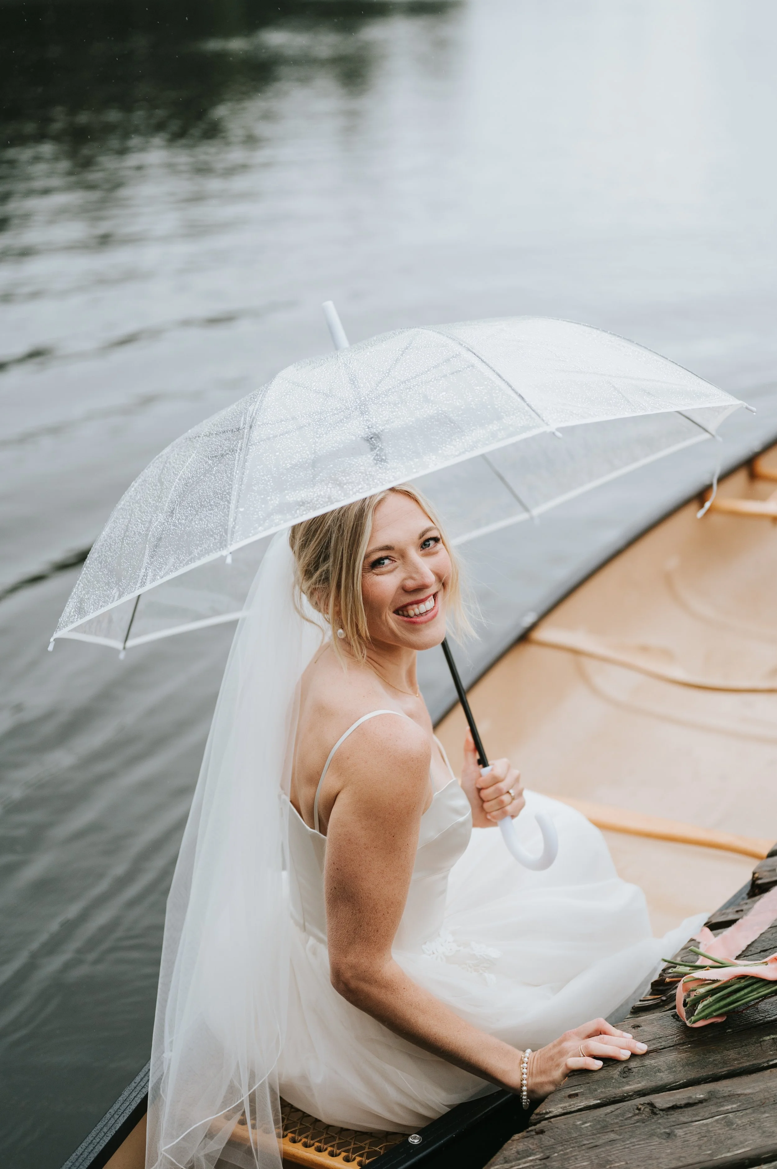 Bride in wedding dress sitting on a boat holding a transparent umbrella with raindrops, smiling at the camera.
