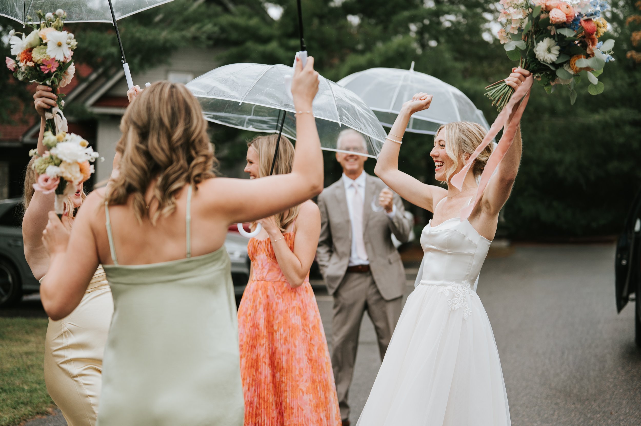 Group of women celebrating outdoors during a wedding, with the bride holding a bouquet and smiling, surrounded by friends holding umbrellas and flowers.