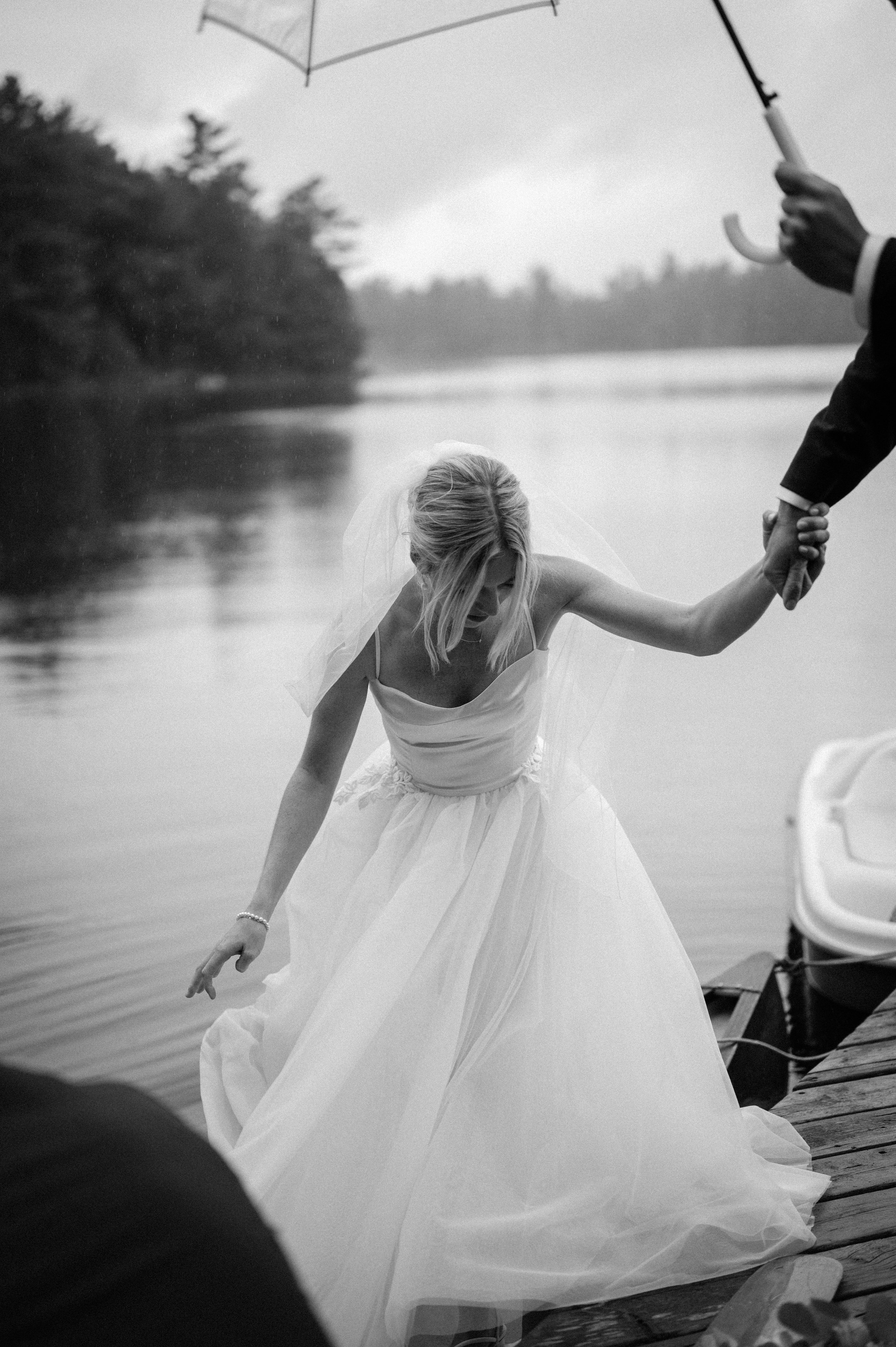 A black and white photo of a bride in a wedding dress stepping out of a boat onto a wooden dock, holding hands with someone holding an umbrella.