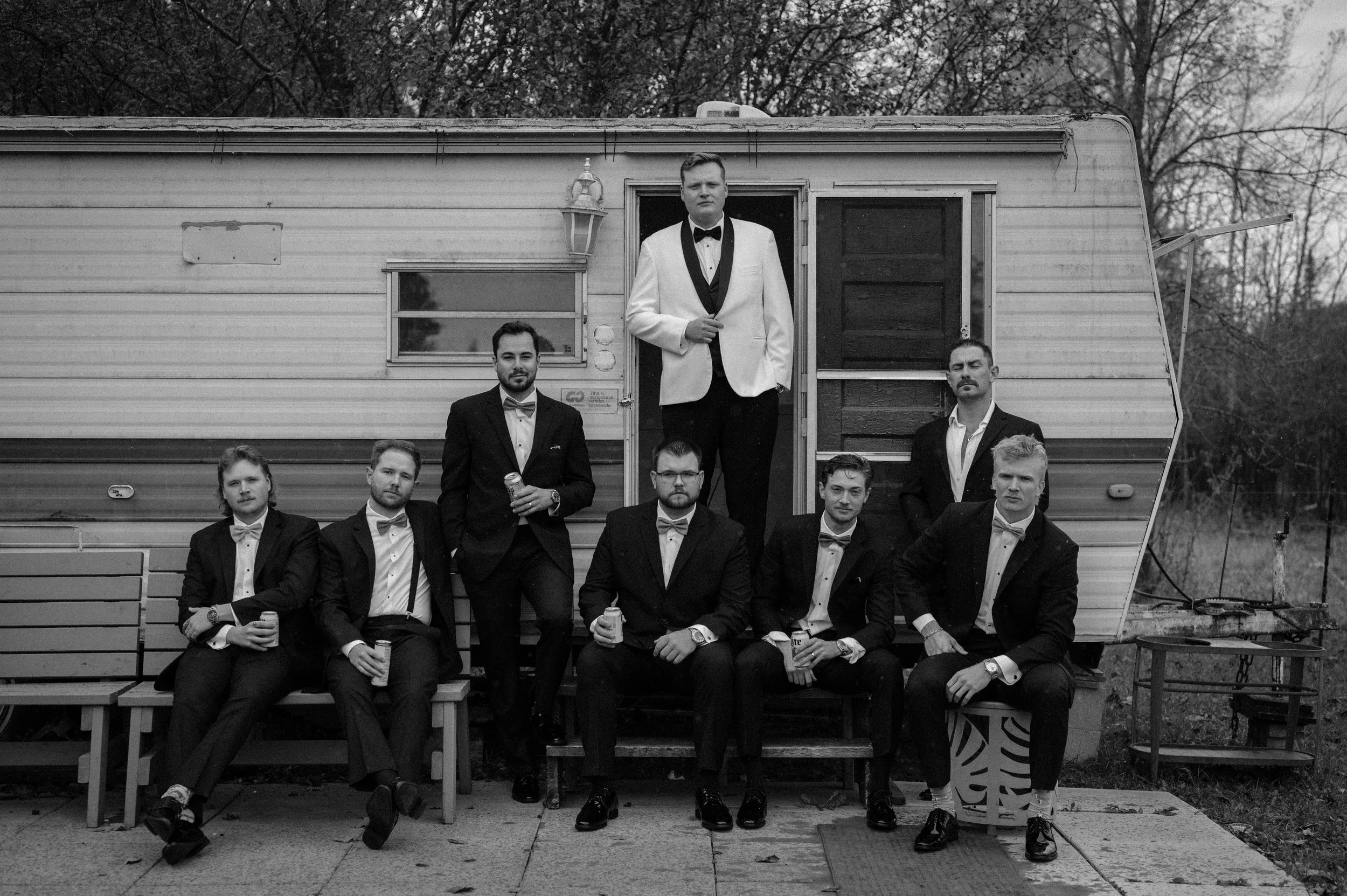 A group of nine men in tuxedos with bow ties, posing in front of a camper. One man is standing in the doorway, while others are sitting or standing nearby, some holding drinks. The scene appears to be outdoors, with trees in the background, and the p