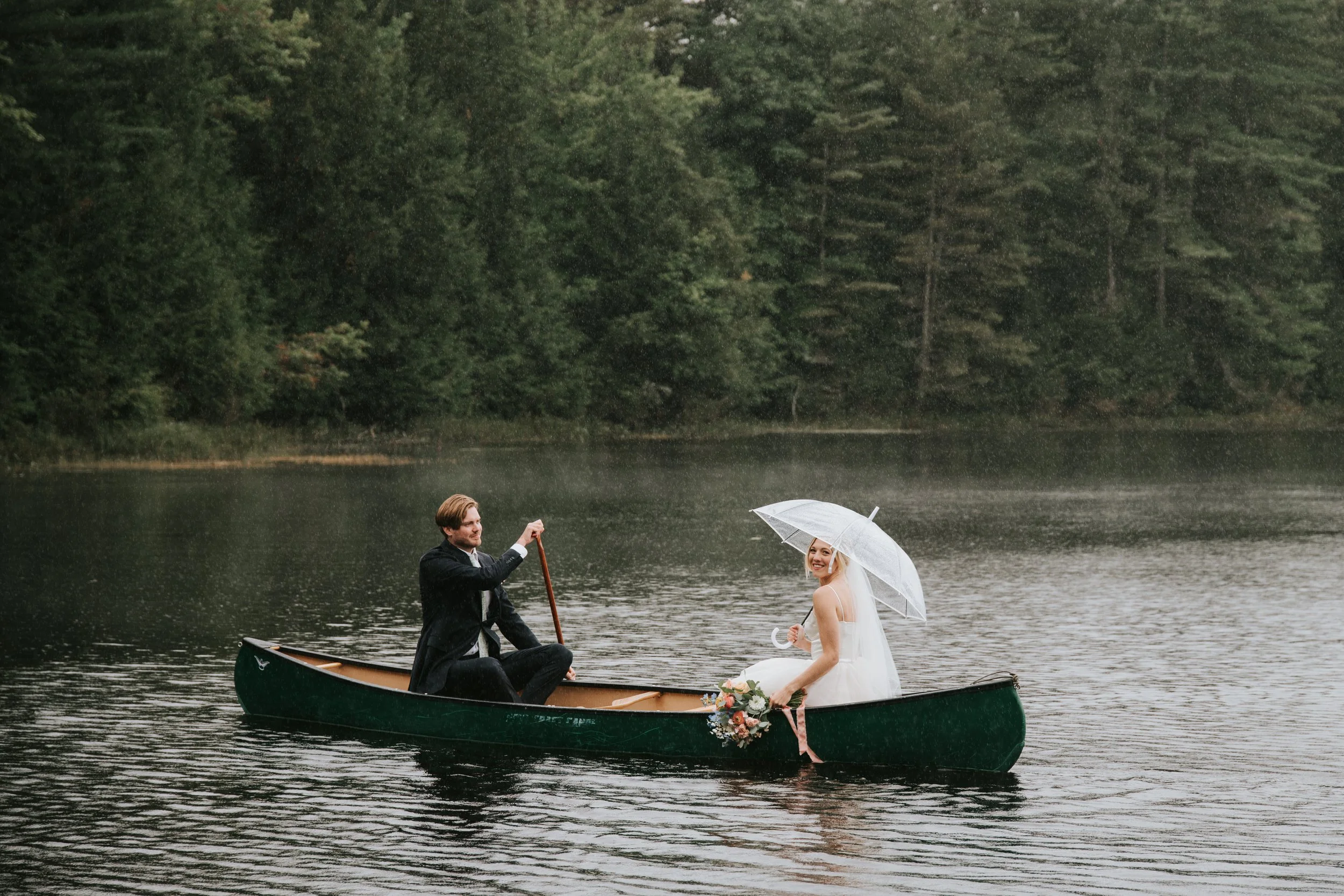 A bride and groom in a canoe on a calm lake, rain falling, surrounded by trees. The bride holds a bouquet of flowers and an umbrella, smiling at the camera; the groom is sitting, holding a paddle.