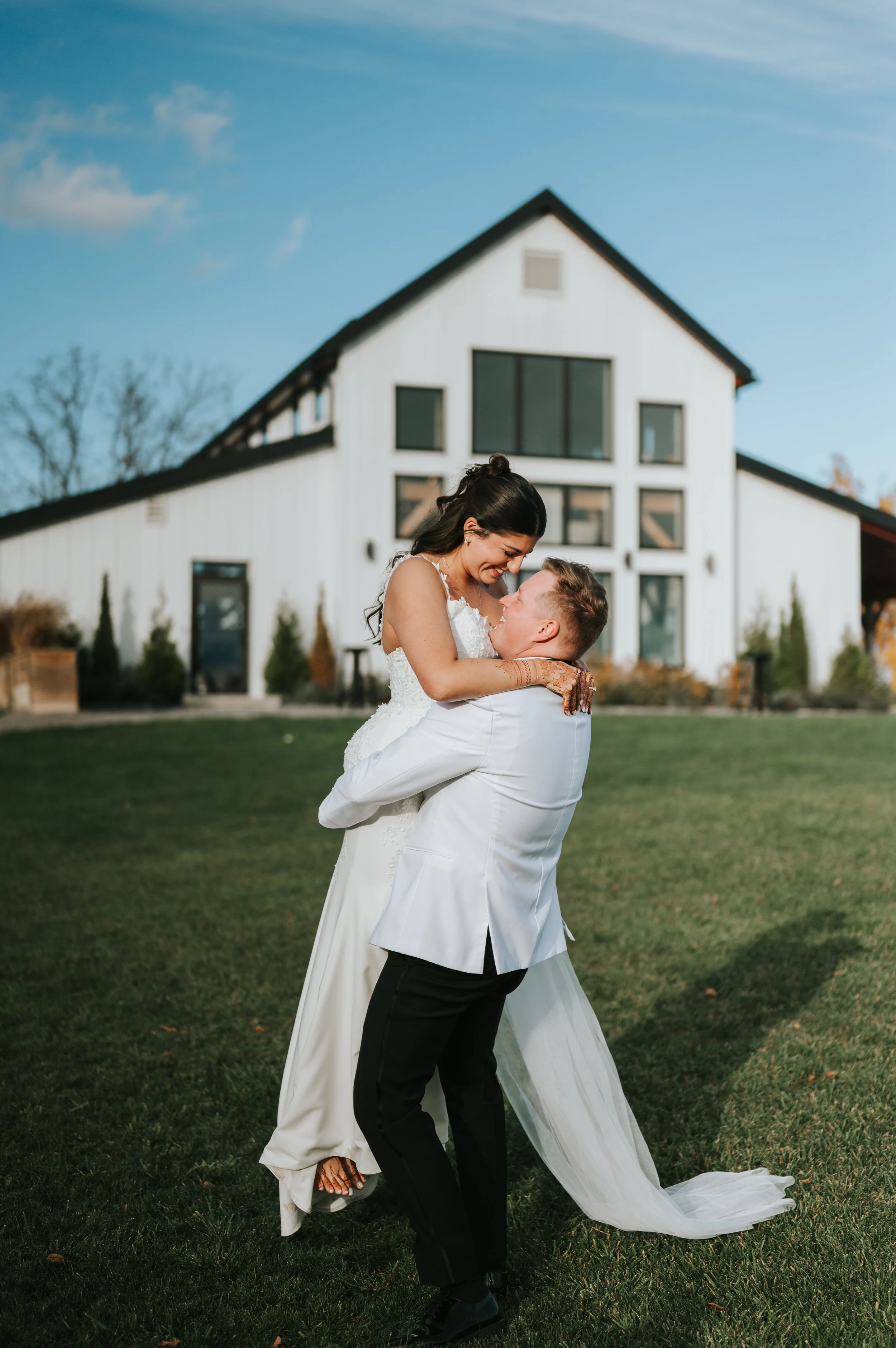 A bride and groom are dancing outdoors in front of a modern white house, with the groom lifting the bride, smiling at each other.