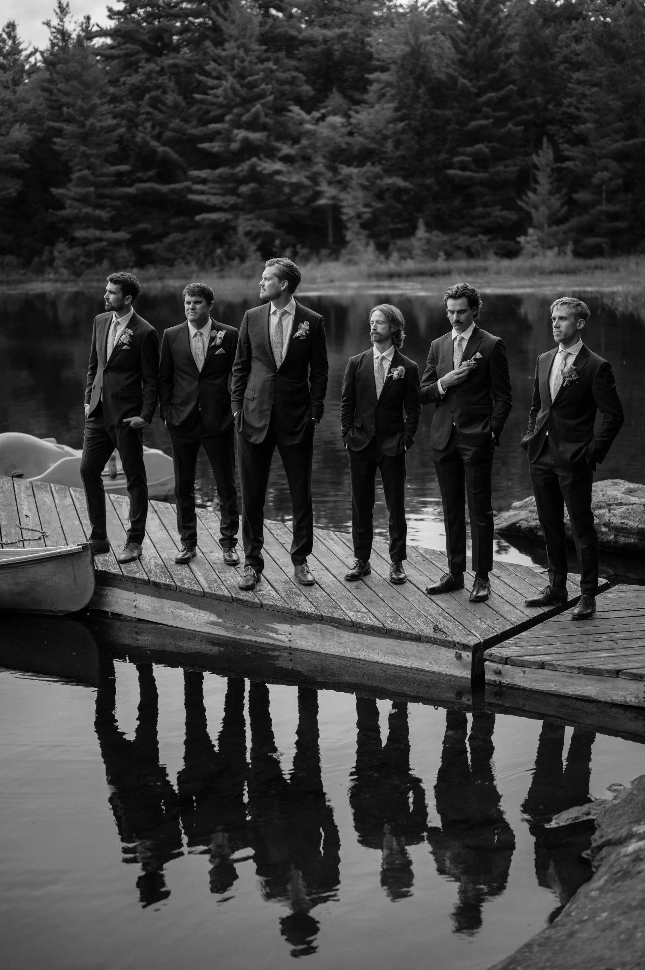Black and white photograph of seven men in suits standing on a wooden dock by a lake with trees in the background, reflecting in the water.