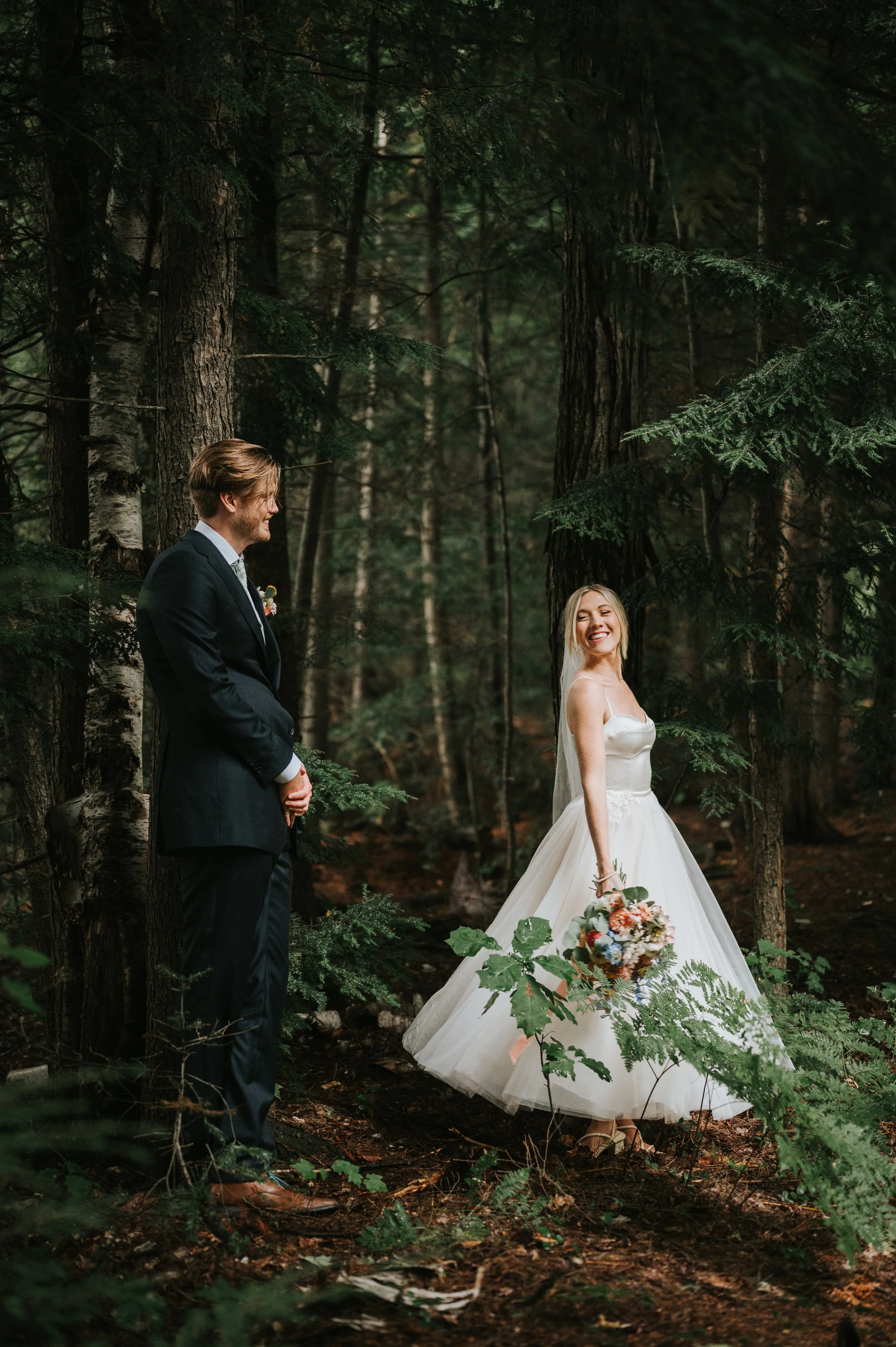 A bride and groom standing in a wooded forest, with the bride smiling and holding a bouquet, and the groom looking affectionately at her.