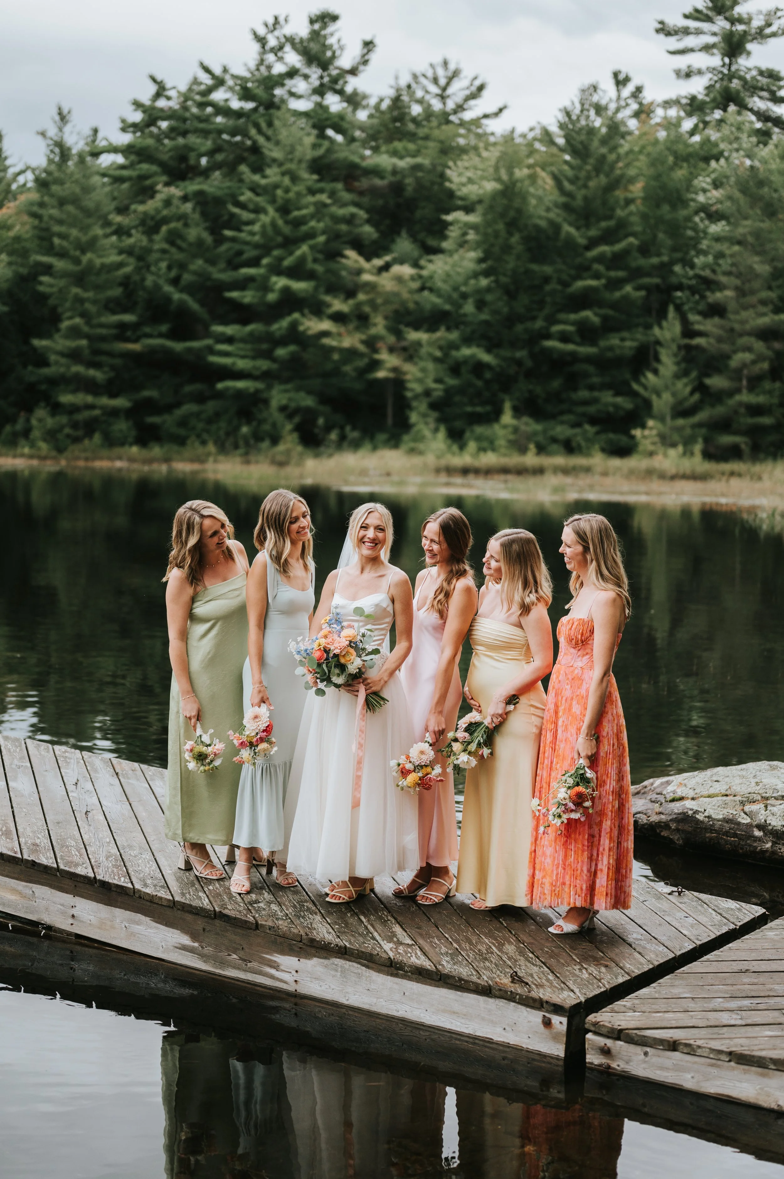A bride in a white wedding dress and veil stands on a wooden dock by a lake, surrounded by five bridesmaids in colorful dresses holding bouquets, all smiling and looking at each other with a forested background.
