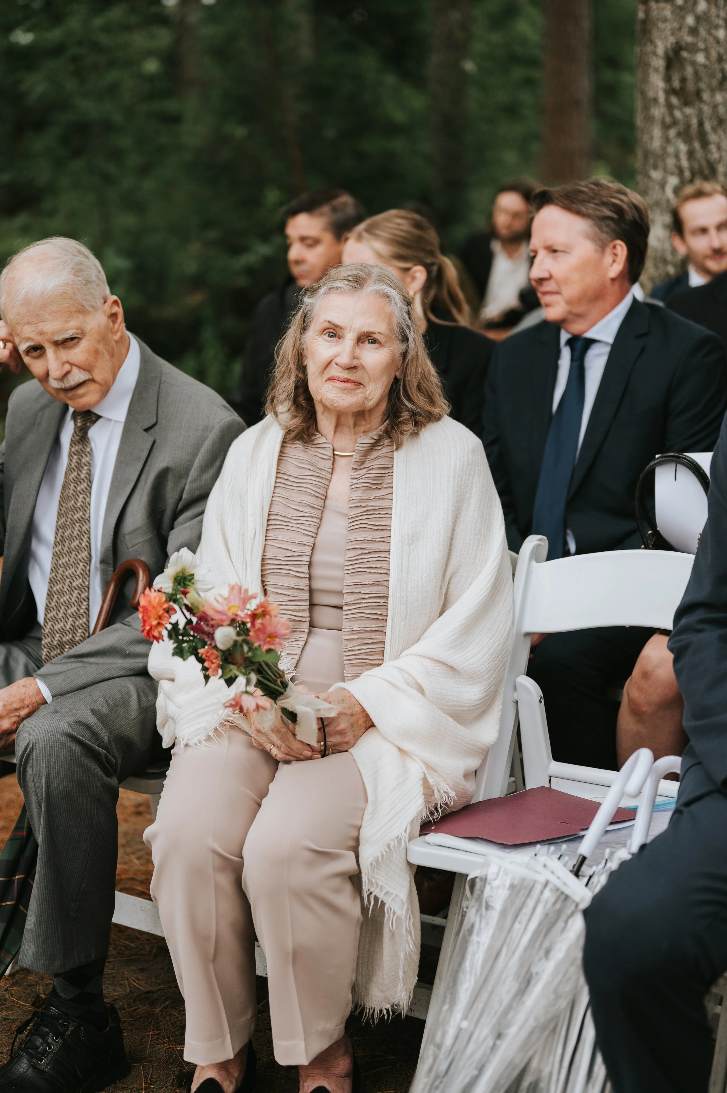 Elderly woman in beige clothing holding a bouquet of pink and white flowers at an outdoor event seated next to a man in a gray suit and tie, with other well-dressed people in the background.