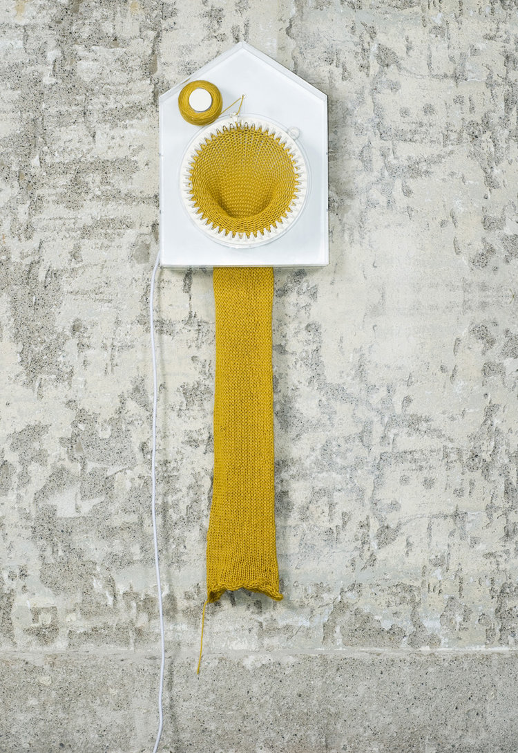 A white clock that is shaped like a house, with a roll of yellow yarn on a thick peg in the top left corner. In the center of the clock is a large circle, with the strands of the yellow scarf that is being knitted beginning on the edge of the circle. The knitted scarf dangles out the back of the clock and downwards