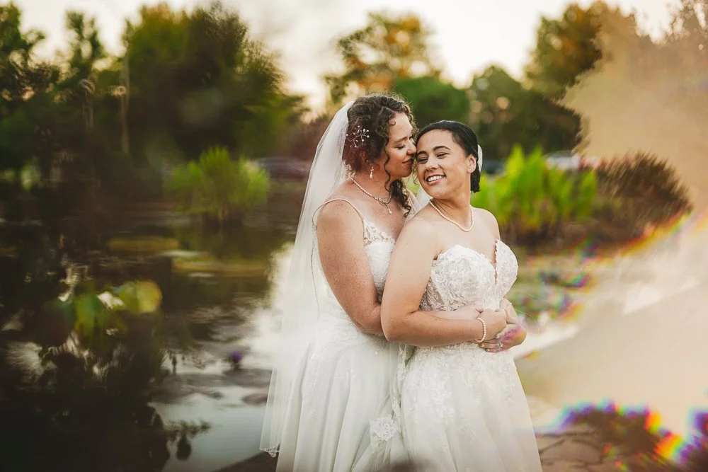 two brides hug, one behind the other, in front of a reflecting pond surrounded by a lush garden