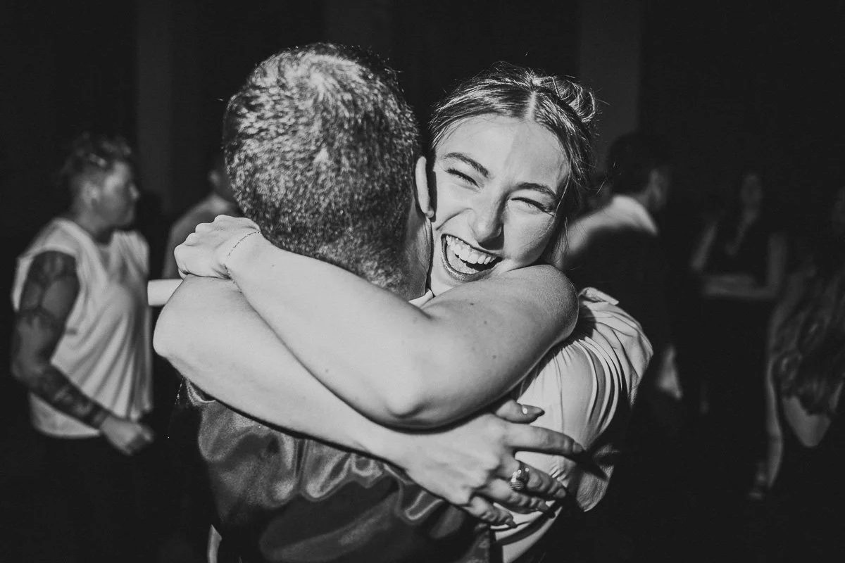 black and white image of a bride hugging a groom on the dance floor. The bride is smiling wide and her arms are fully wrapped around his head.
