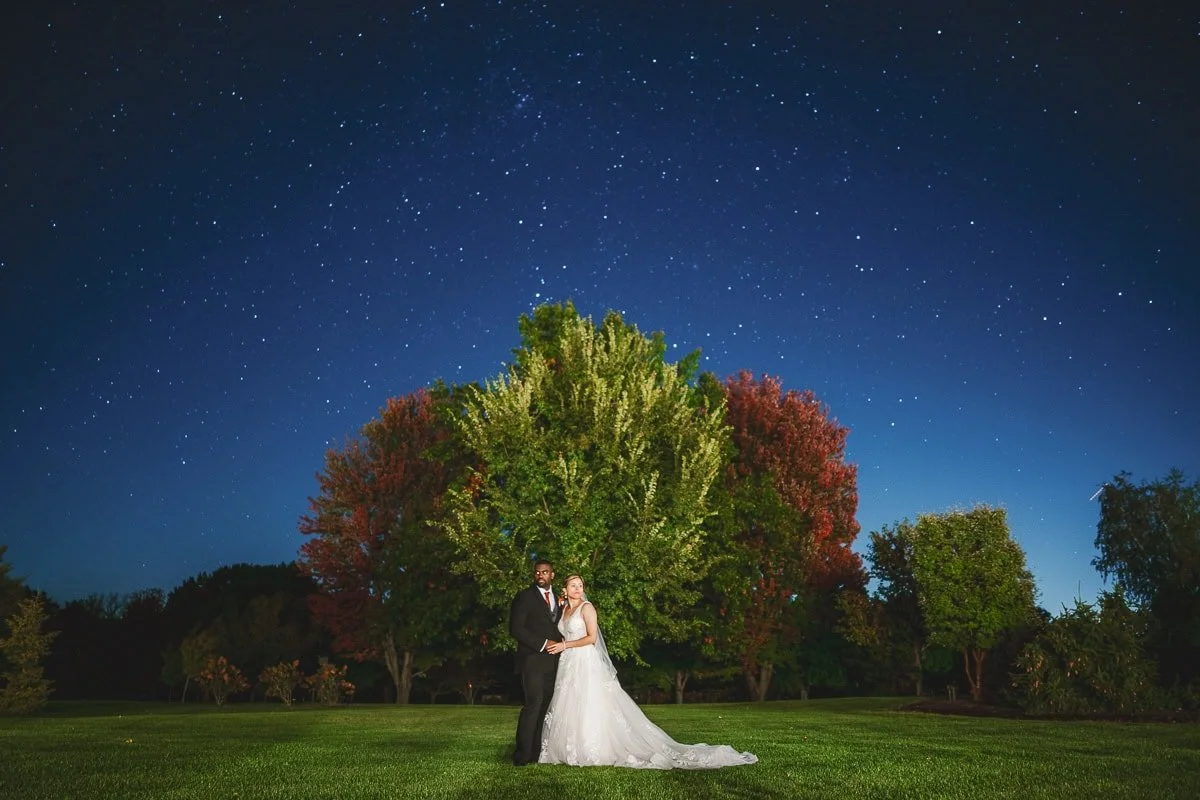 couple stands in a clearing at night looking opposite directions, trees with fall colors behind them, and a starry night sky above them