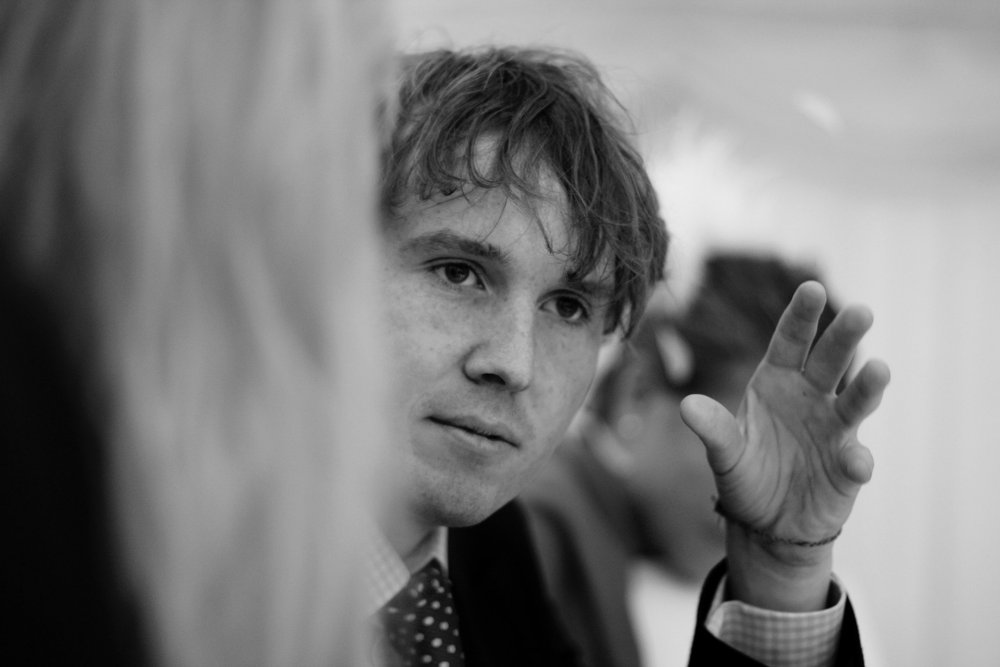 A young man with messy hair and freckles, making a gesture with his hand while talking.