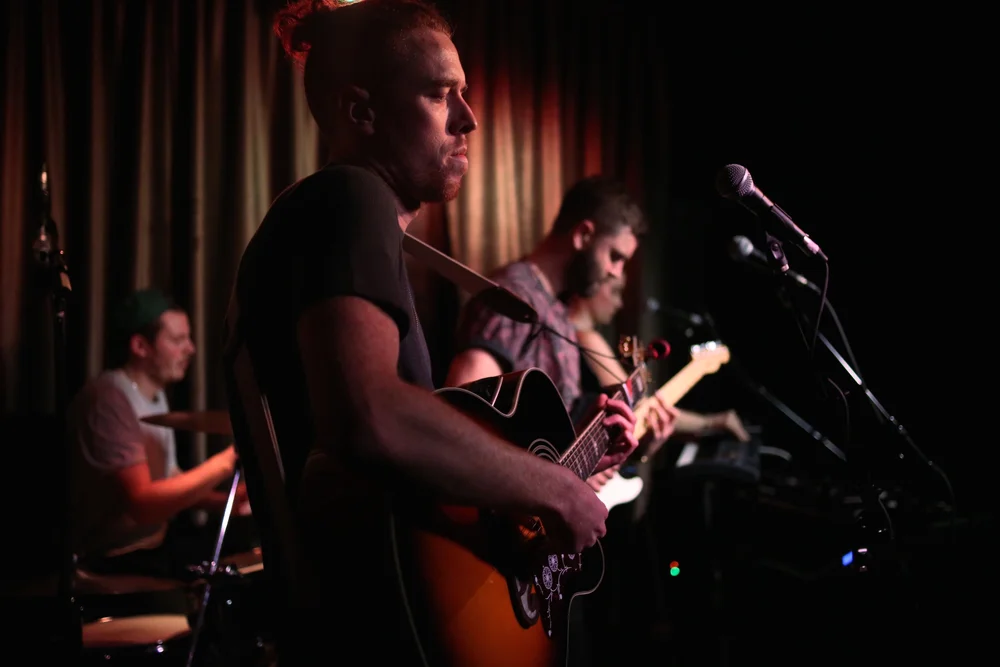 A band performing live on stage, with one musician playing an acoustic guitar in the foreground and other members playing electric instruments in the background, stage lighting creating a warm ambiance.
