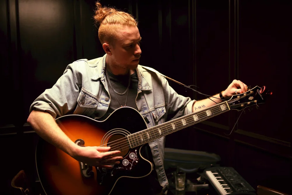 A young man with blonde hair playing an acoustic guitar in a dark room.