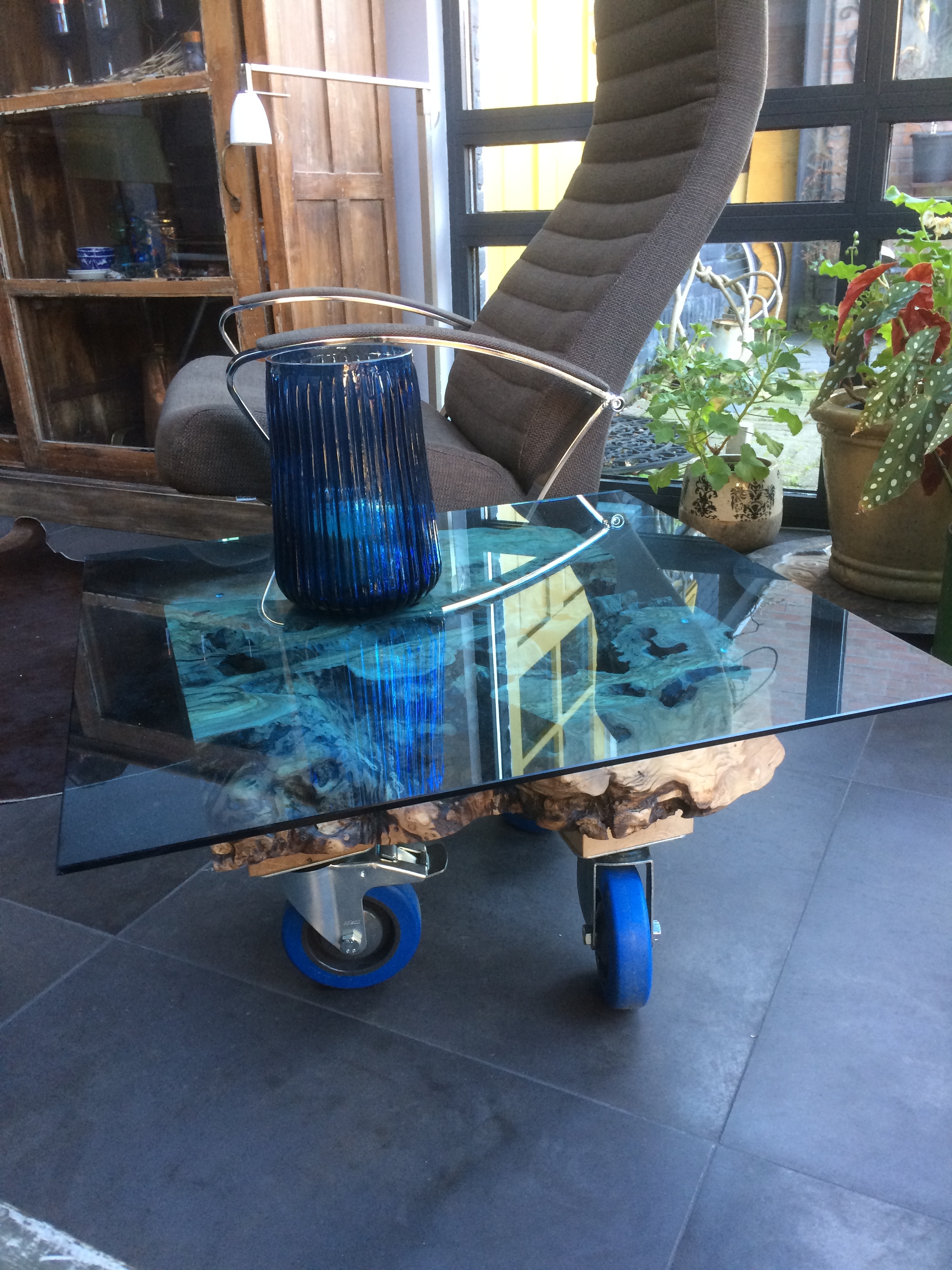 Olijfhouten Salontafel met blauwe glasplaat, het hout is 400 jaar oud en de tafel heeft drie industriële wielen. Afmeting 60 x 60 cm.