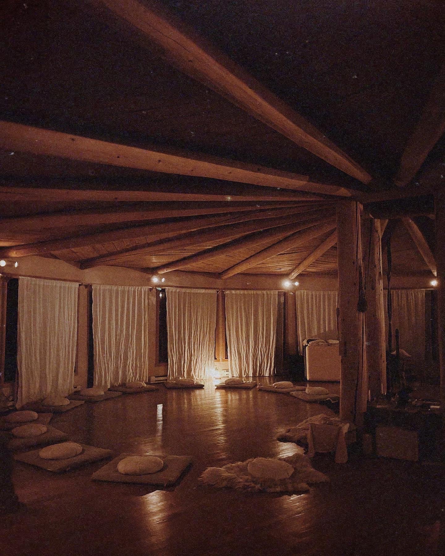 Dimly lit meditation room with round arrangement of cushions on wooden floor, surrounded by draped curtains and wooden beams overhead.