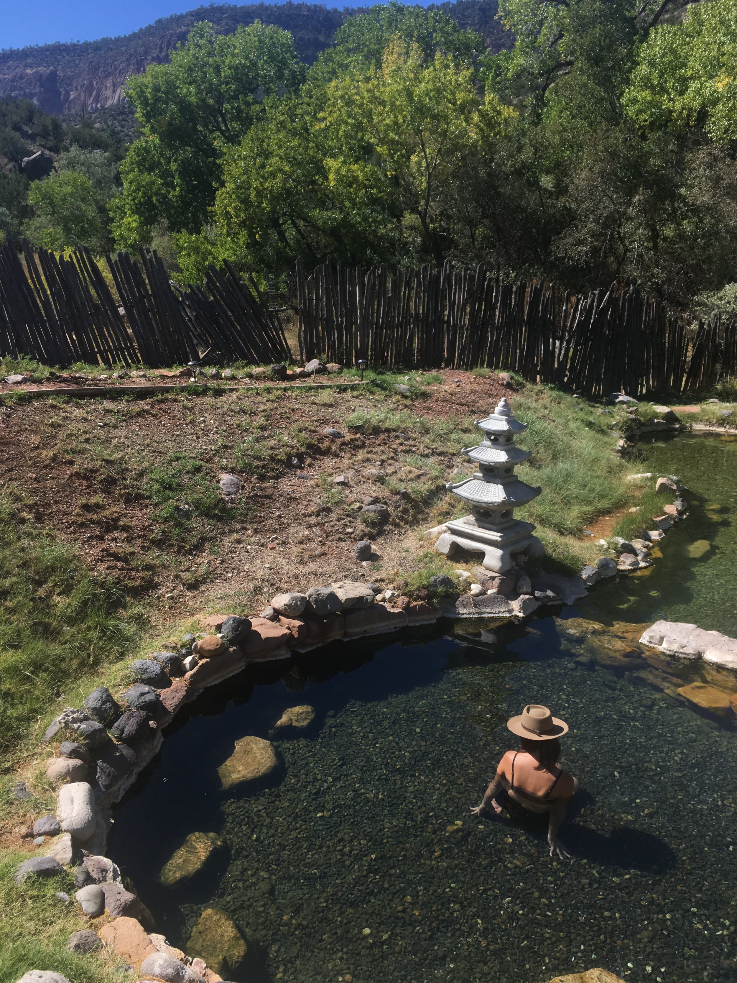 Person sitting in a small stone-lined pond in a natural setting with trees, wooden fence, and decorative stone sculpture.