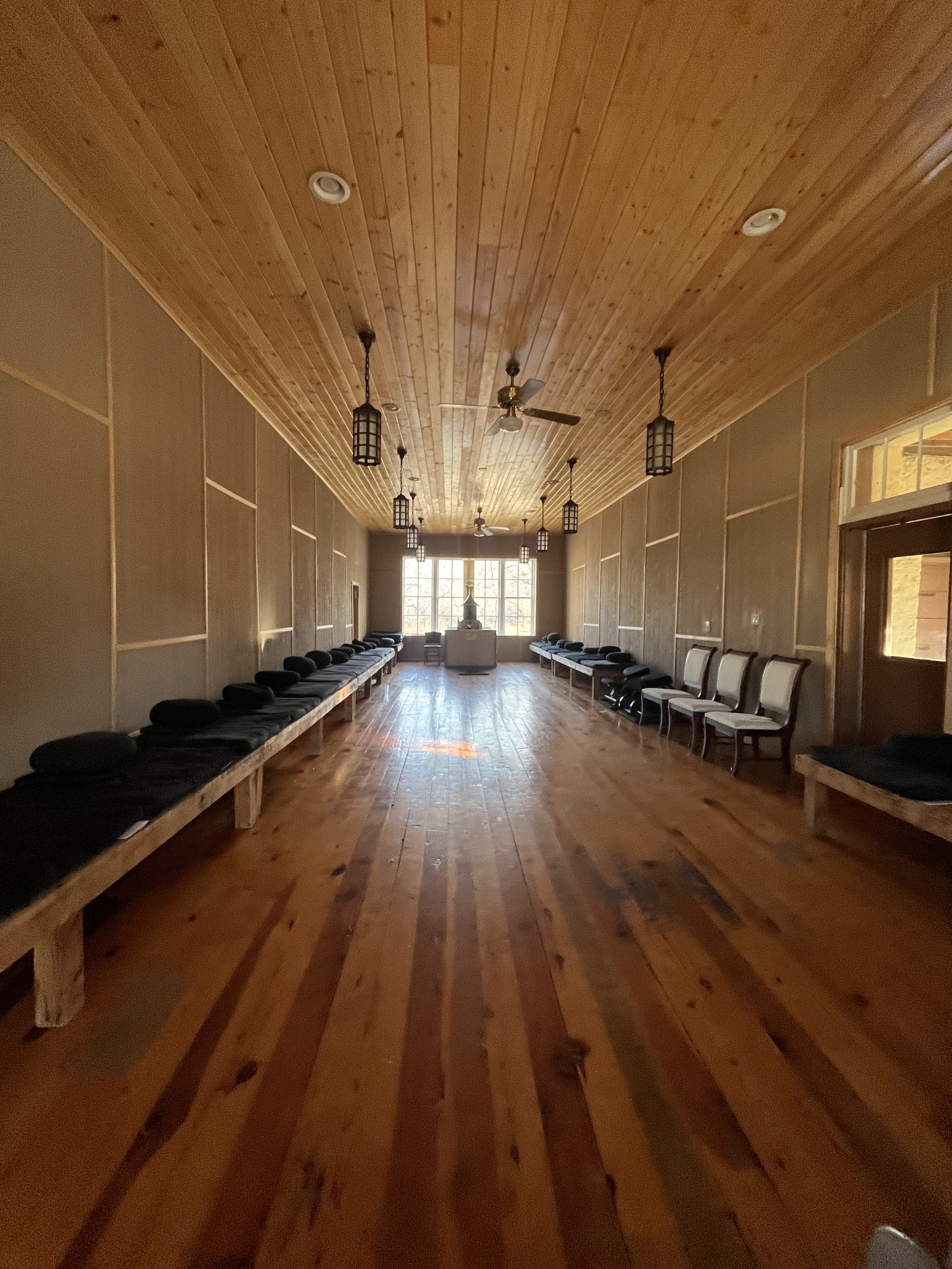 Wooden meditation hall with bench seating and cushions, hanging lanterns, and a ceiling fan.