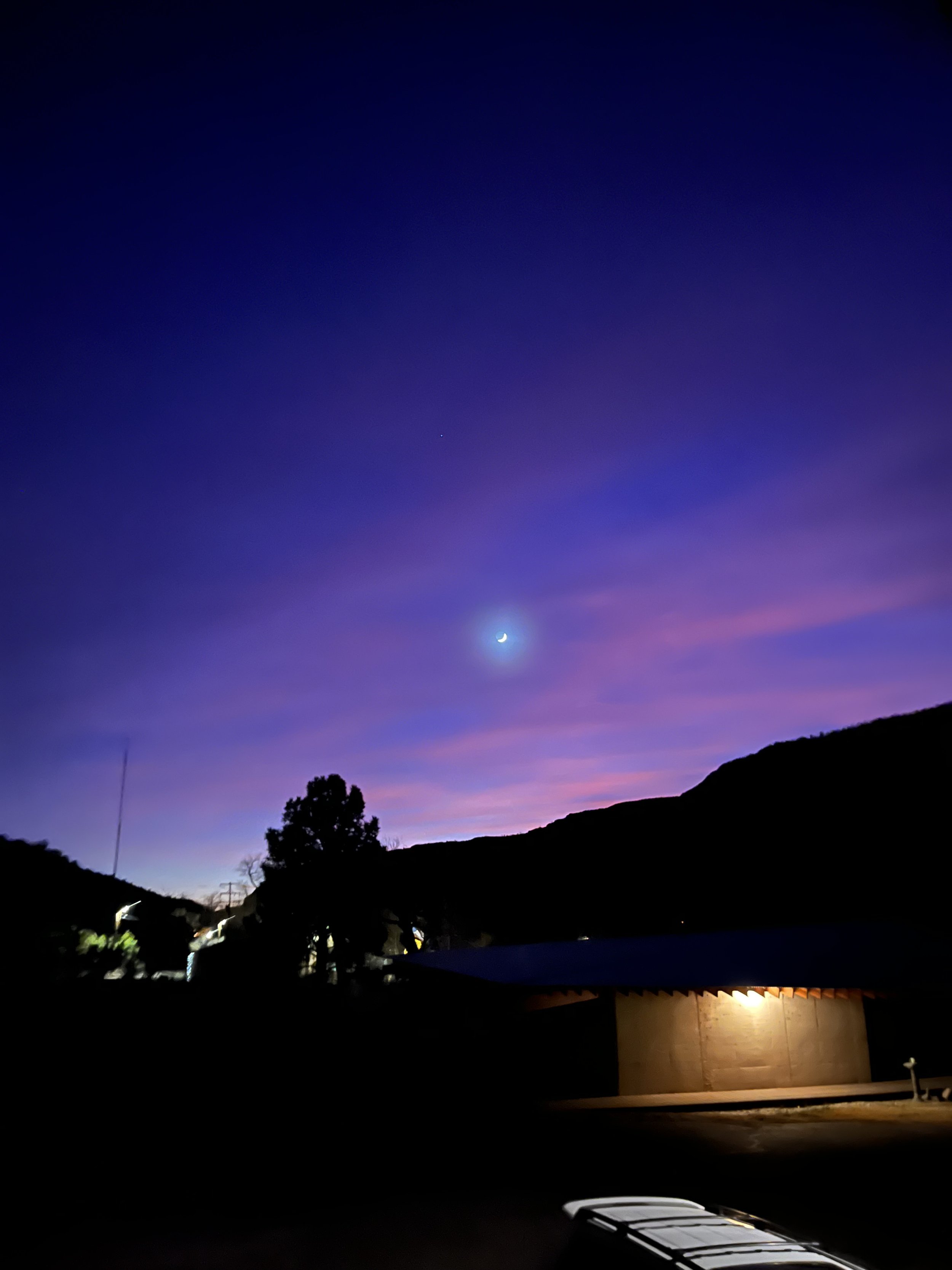 Night sky with crescent moon and glowing horizon over silhouetted landscape and building.