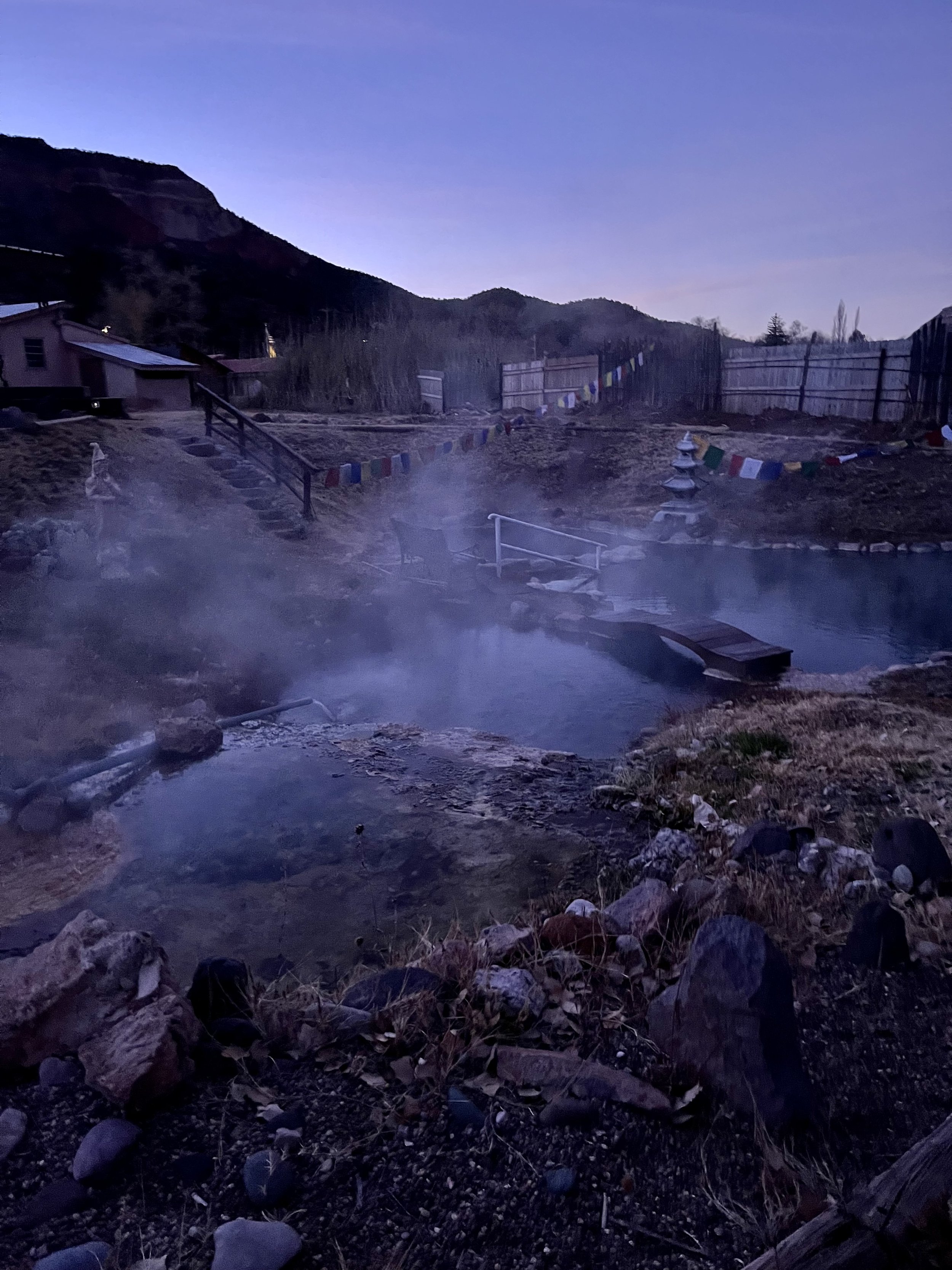 A hot spring surrounded by rocks and vegetation at dusk, with steam rising from the water. In the background, there is a wooden fence decorated with colorful prayer flags and stairs leading to a small building. Hills are silhouetted against the evening sky.