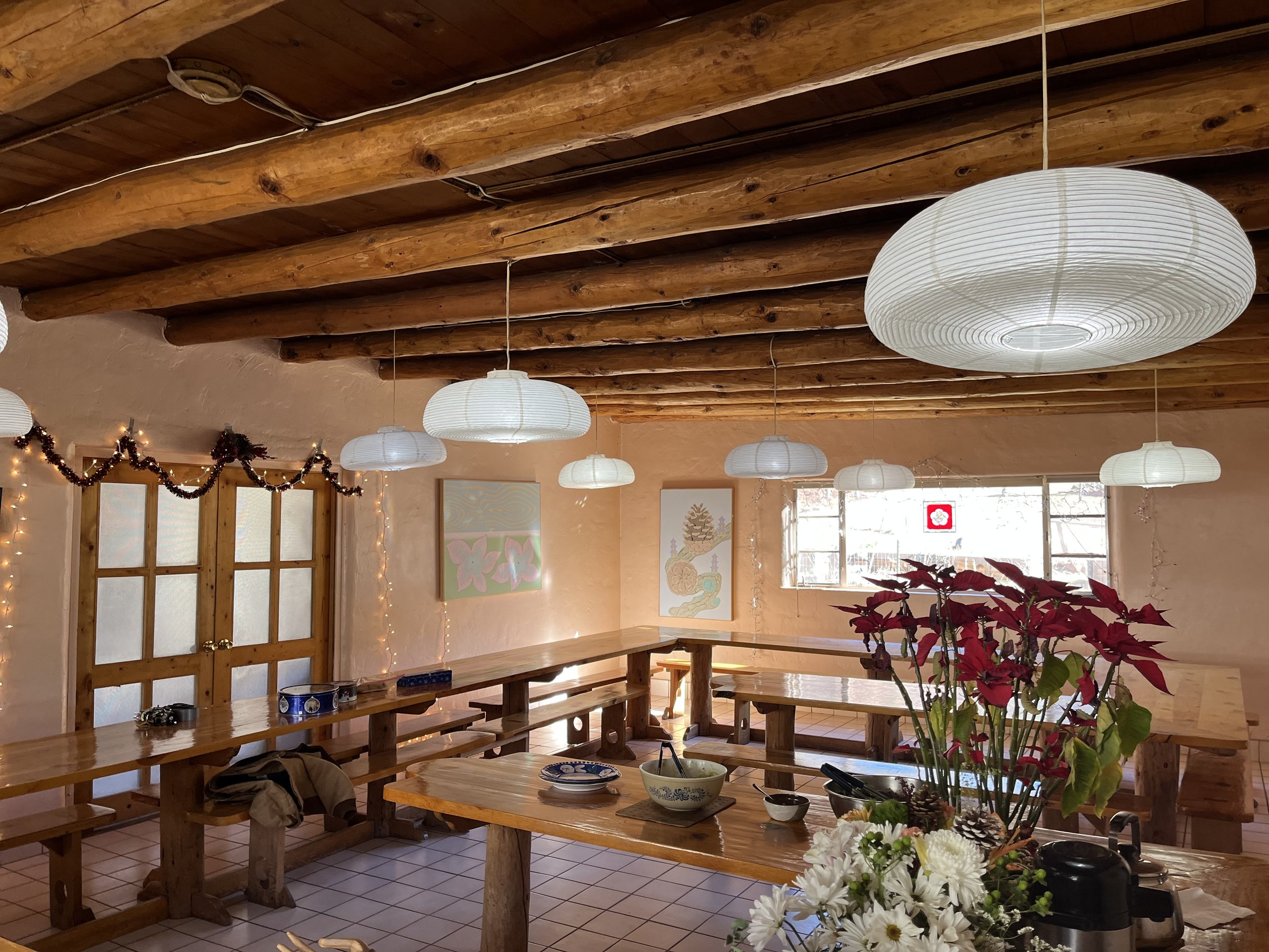Rustic dining area with wooden ceiling beams, long wooden tables, and benches. Decor includes hanging paper lanterns, string lights, poinsettias, and daisies. Artwork and frosted glass double doors are also visible.