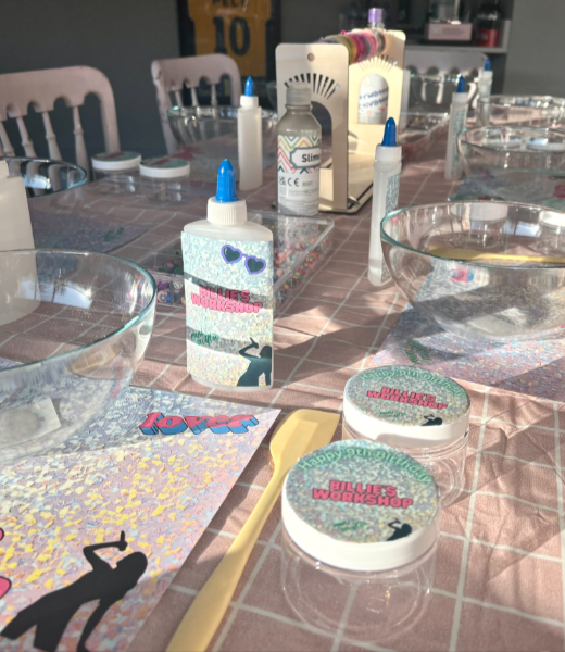 Table set up for slime crafting activity with bottles of slime, paintbrush, and decorative papers, on a pink checkered tablecloth.