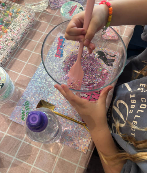 Child mixing colorful glitter and beads in a clear bowl with a pink spatula, surrounded by craft supplies and decorated paper on a checkered table.