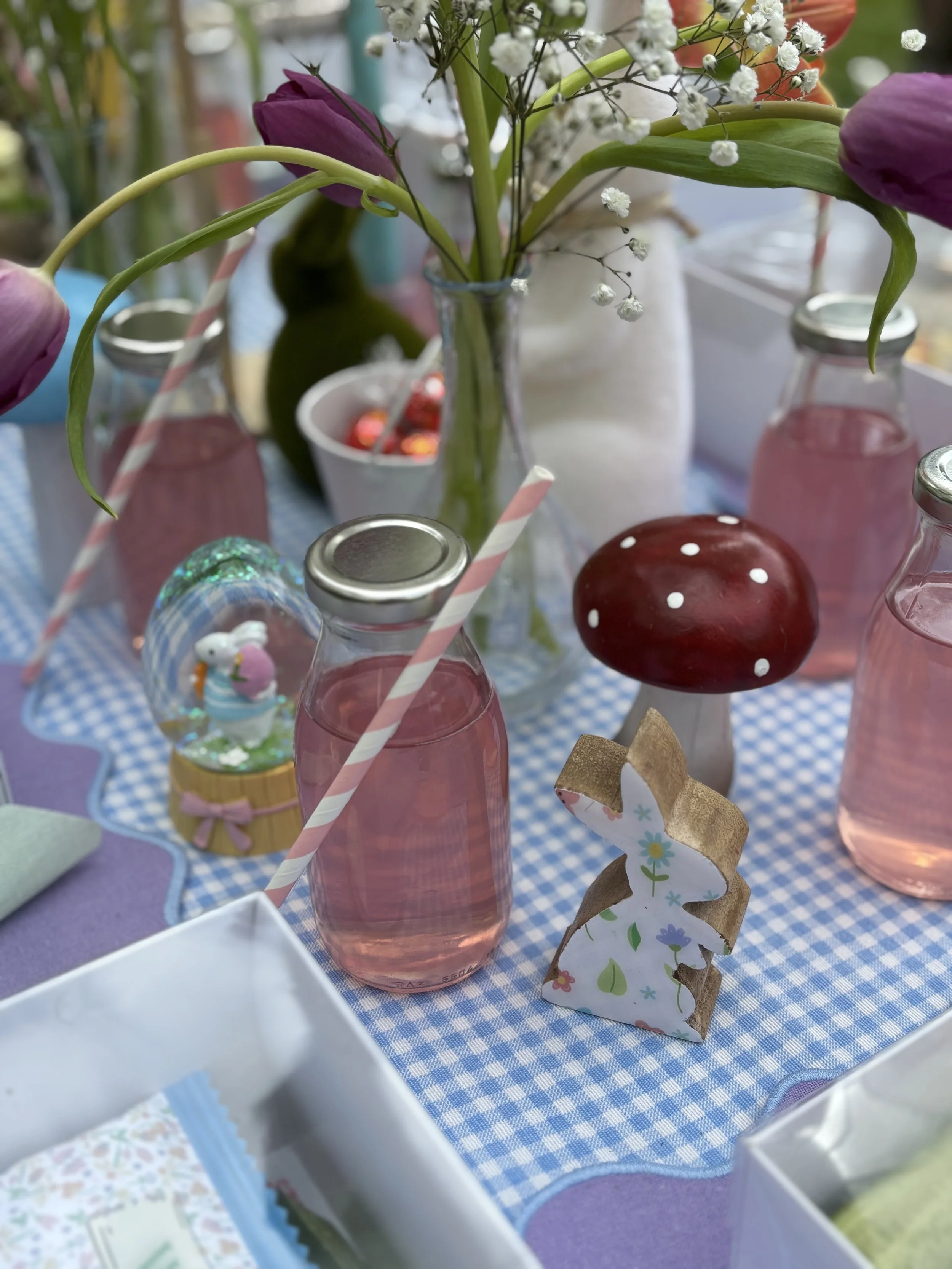 A table decorated with a blue and white checkered tablecloth featuring pink bottles with striped straws, a small mushroom-shaped figure, a decorative bunny with a floral pattern, a snow globe with a bunny figure, and a flower arrangement in a glass vase. The flowers include purple tulips and white baby's breath.