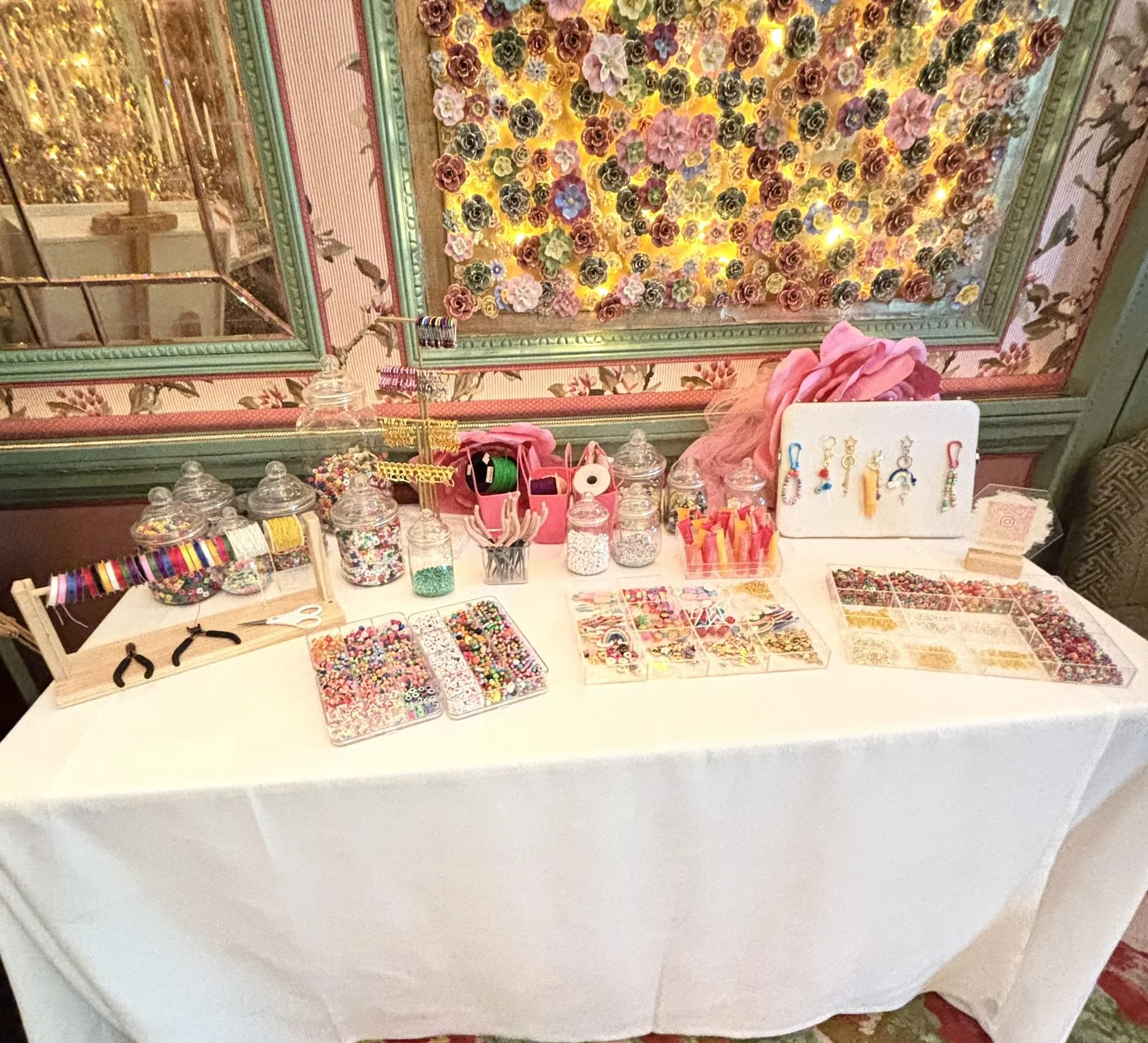 A display table with jewelry-making supplies, including colorful beads, thread, and tools, set against a floral wall with pink, yellow, and purple flowers.