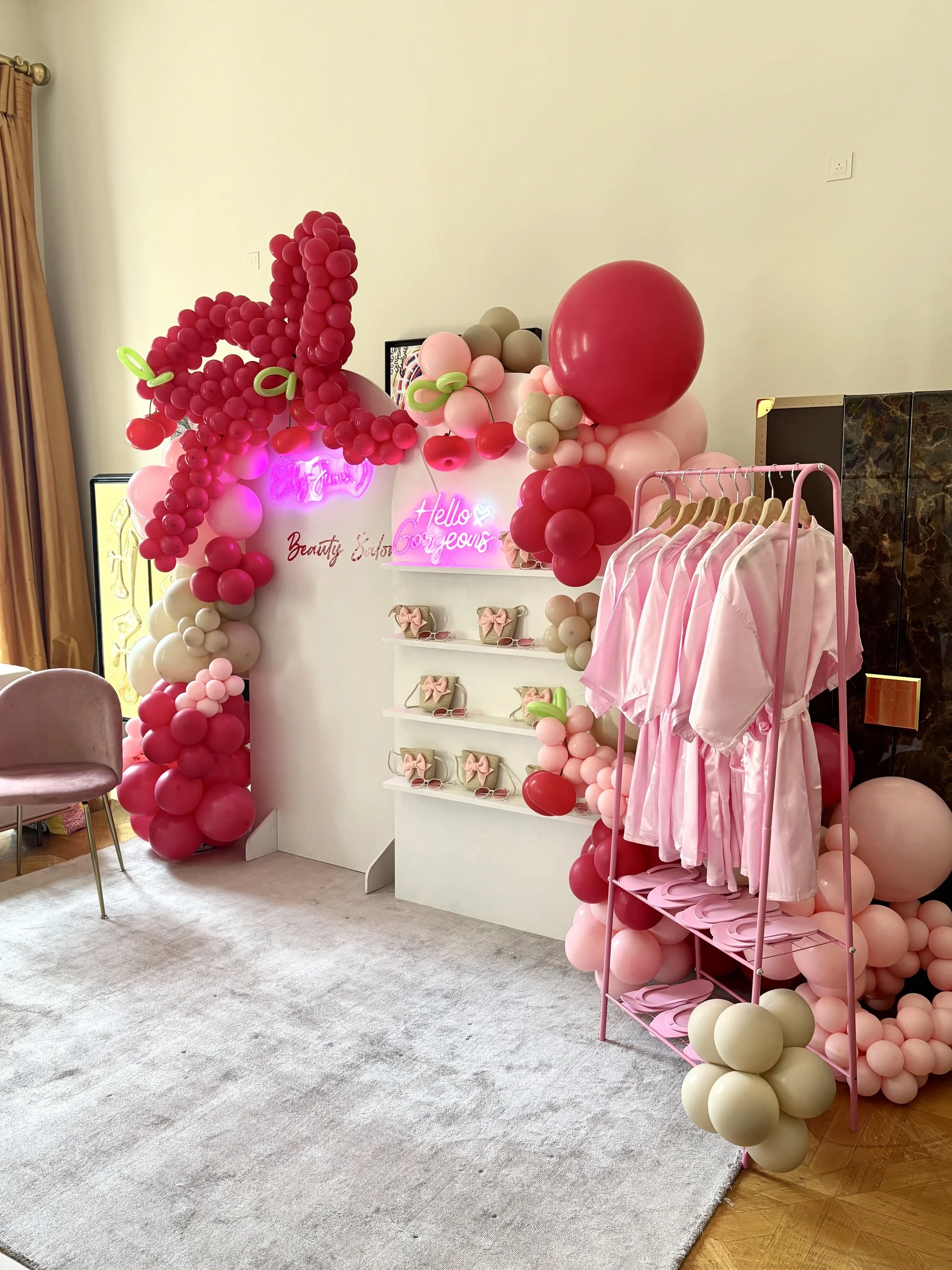 Pink and red balloon arch with neon signs, pink dresses on a clothing rack, and shelves of gift boxes, creating a festive or celebratory setup.