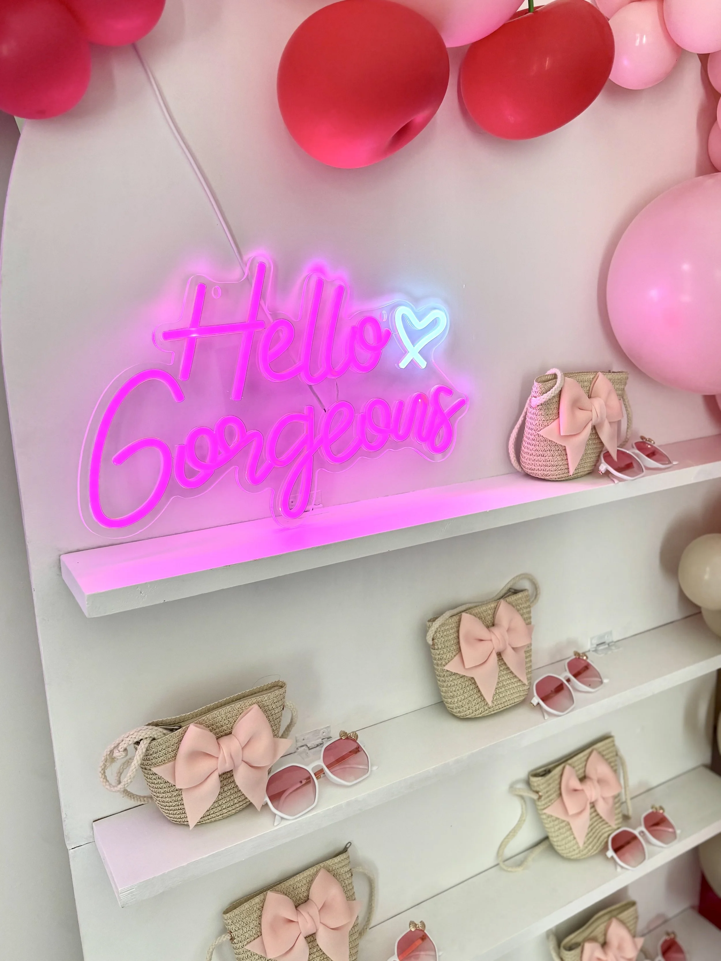 Colorful display with pink balloons, neon sign saying "Hello Gorgeous" with a heart, and small pink and beige bags with bows on storeshelves.