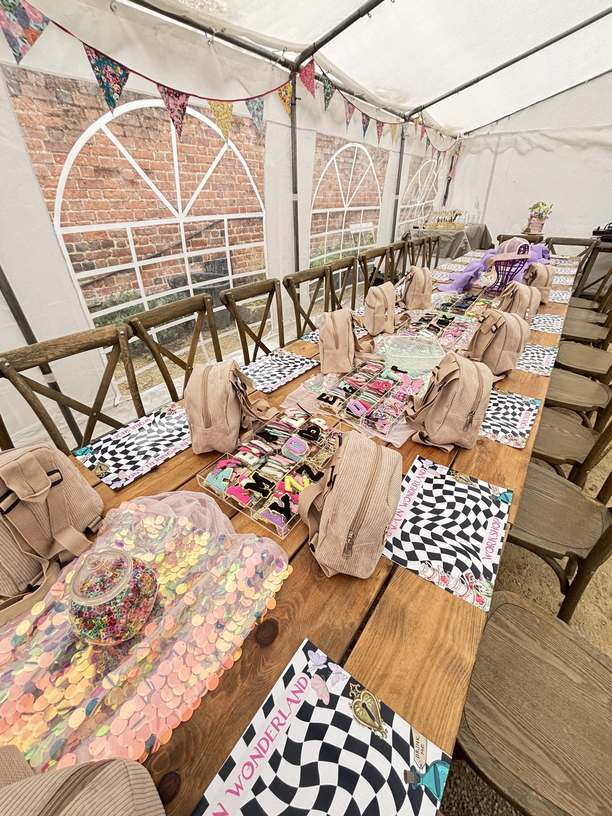 Decorated outdoor event space with a long wooden table set with backpacks, small toys, and party favors, featuring colorful bunting and black-and-white checkered themed place mats.