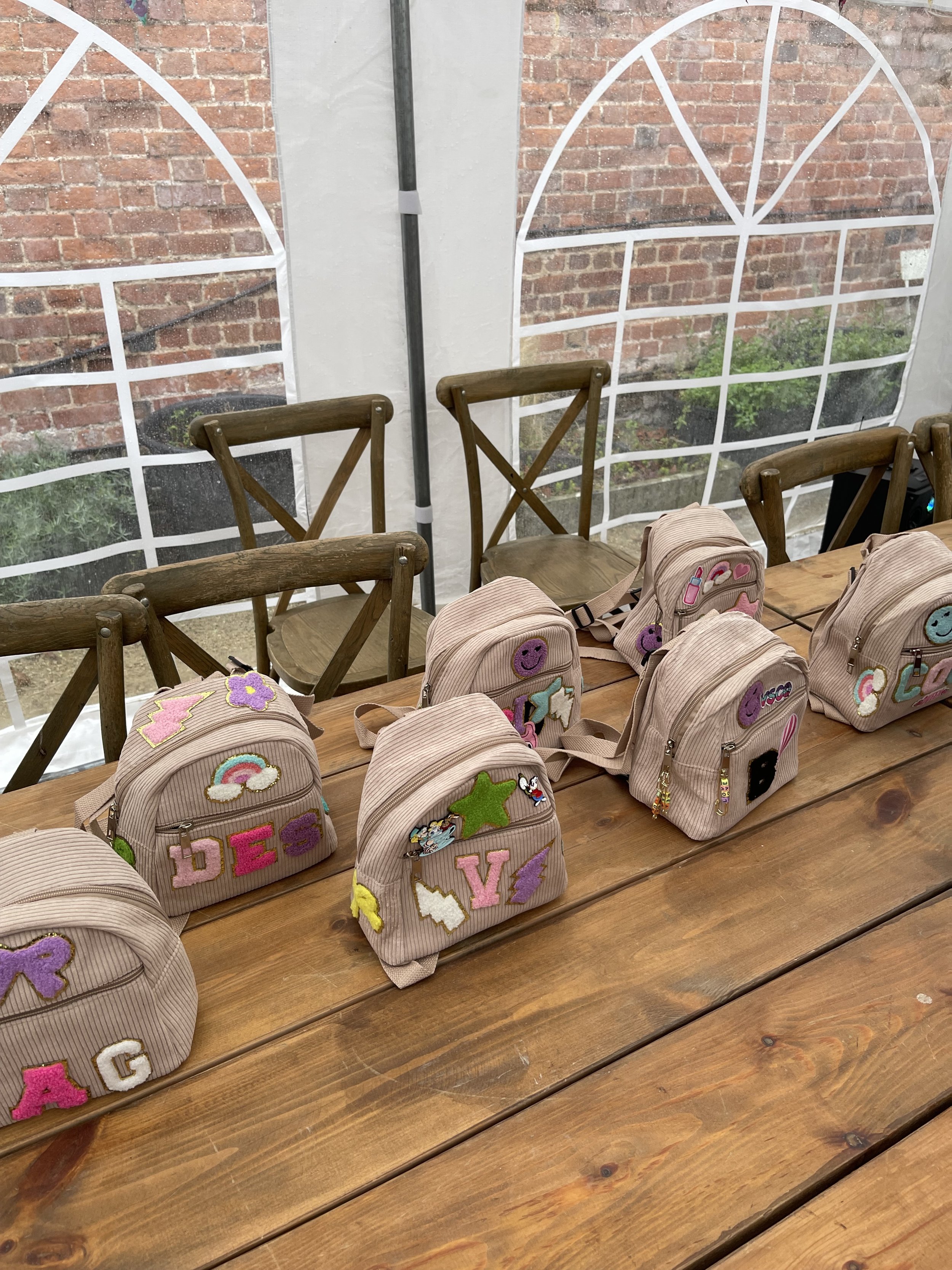 Multiple small backpacks with colorful patches placed on a wooden table inside a tent with transparent windows showing brick wall outside.