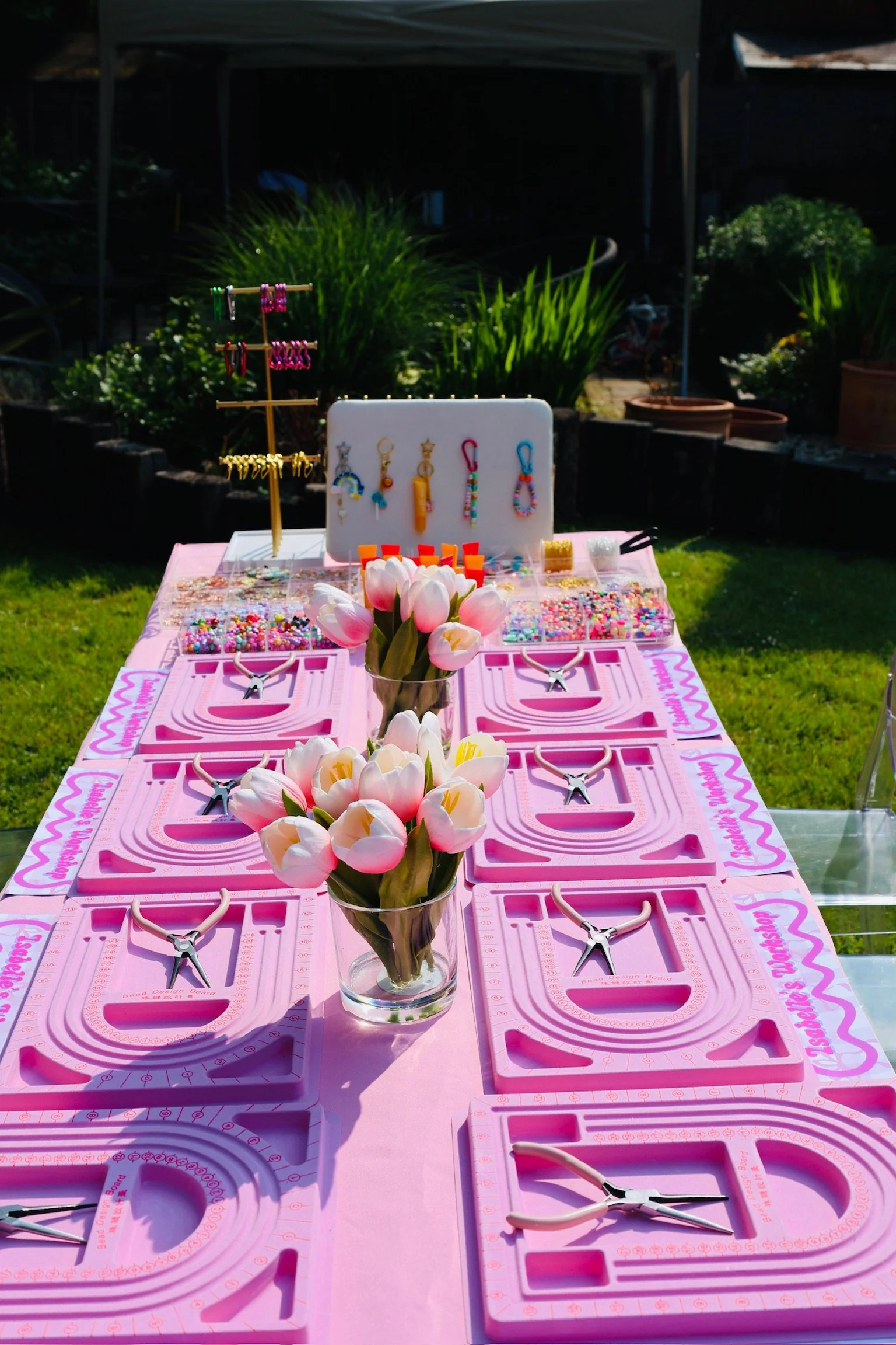 A pink-themed craft table set up outdoors with floral decorations, jewelry-making supplies, and pink jewelry display boards, with plants and a garden background.