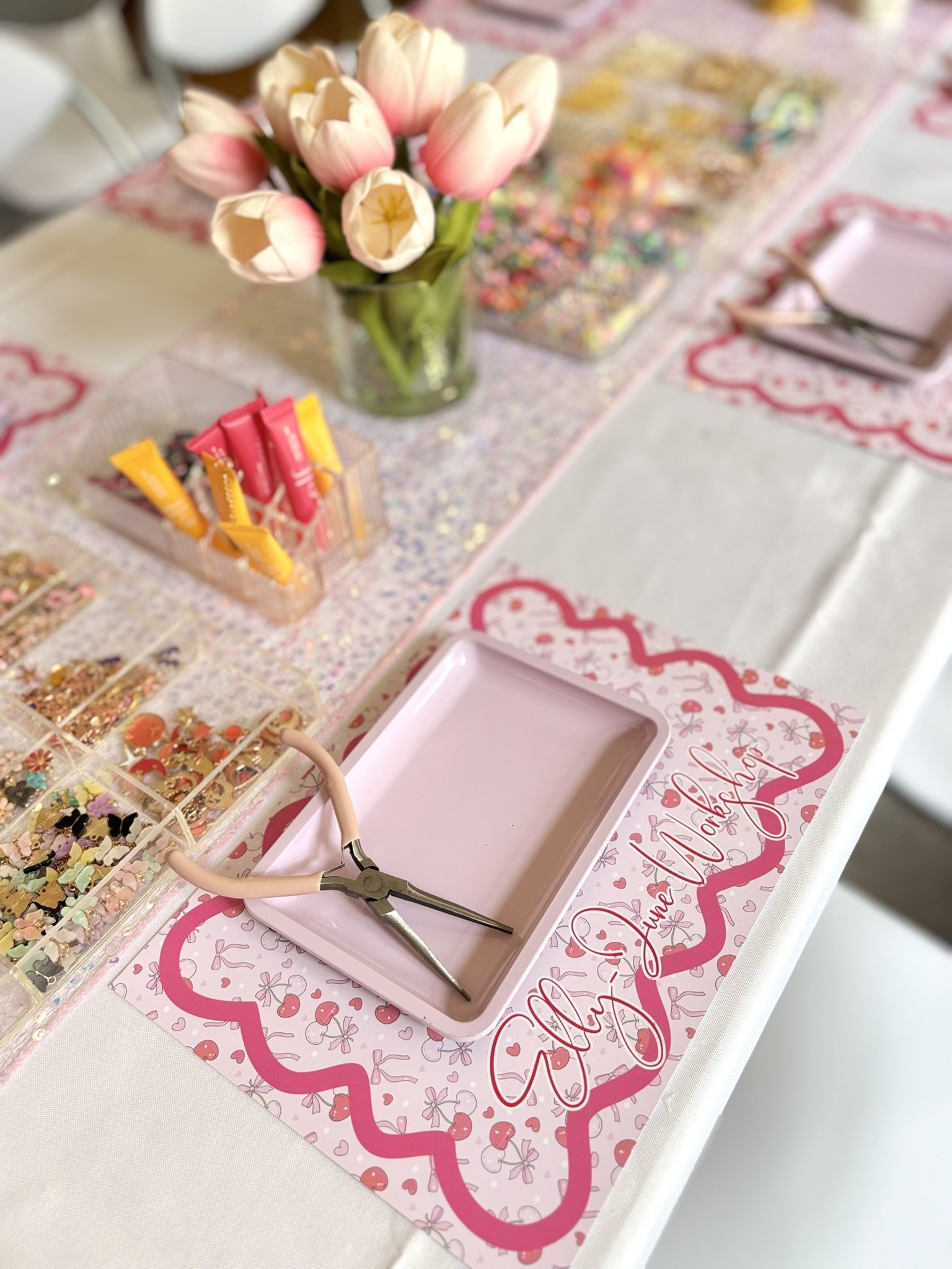 A table decorated with pink and white tulips in a glass vase, colorful sprinkles, and candy. There are pink placemats with a cherry design for a bridal or baby shower, and a jewelry tool laying on one of the placemats.