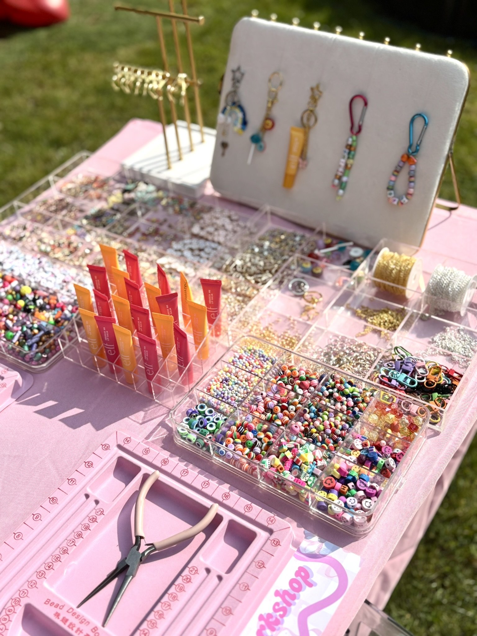 Jewelry-making supplies at a craft booth, including colorful beads, charm clips, and tools on a pink tablecloth.