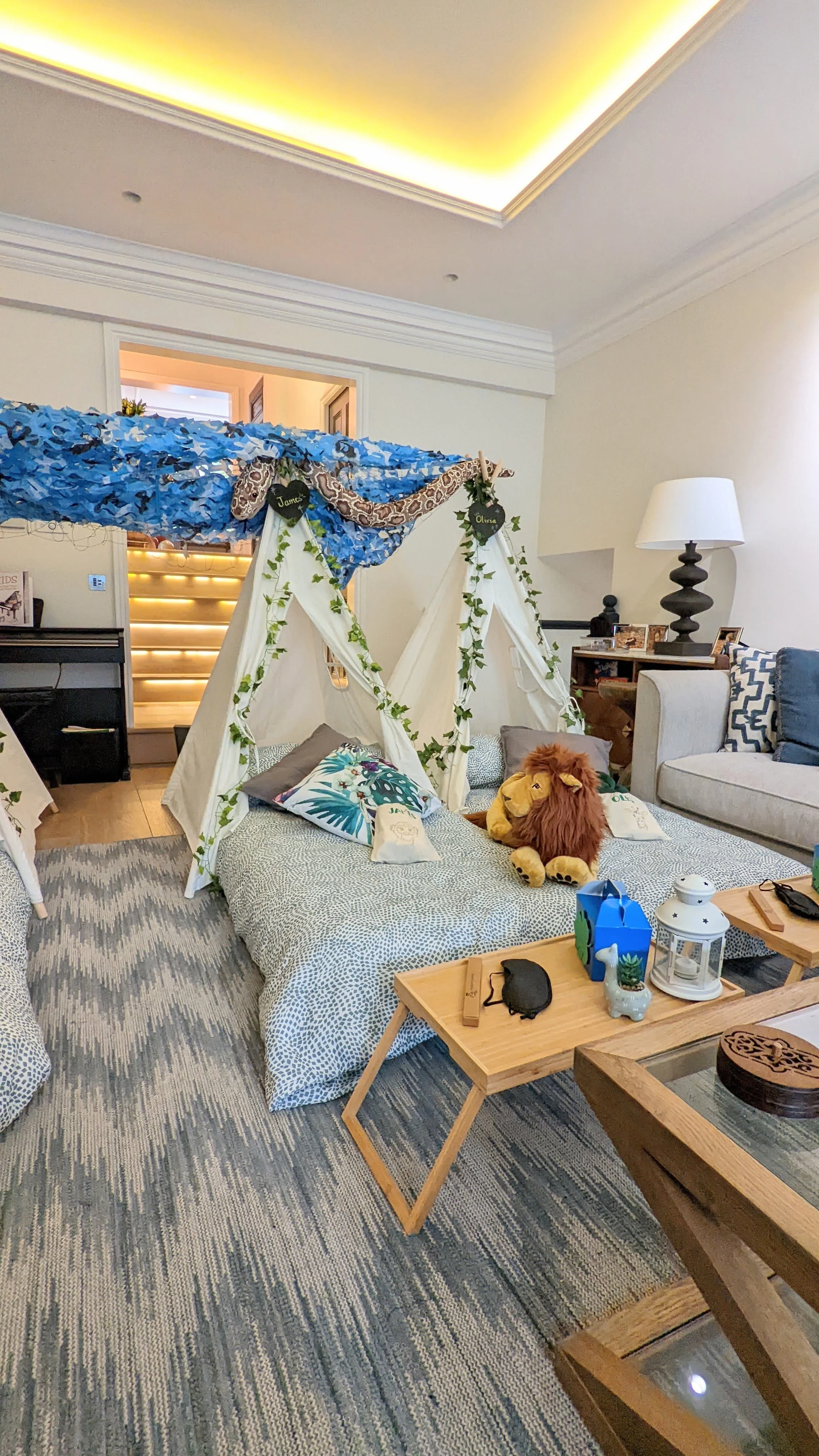 Indoor scene with a small children's play tent set up on the floor, decorated with green ivy and a blue canopy overhead. Inside the tent, there are plush toys, including a lion, and pillows. The room contains furniture such as a side table with items, a sofa with cushions, and a lamp. The flooring is covered with a textured rug.