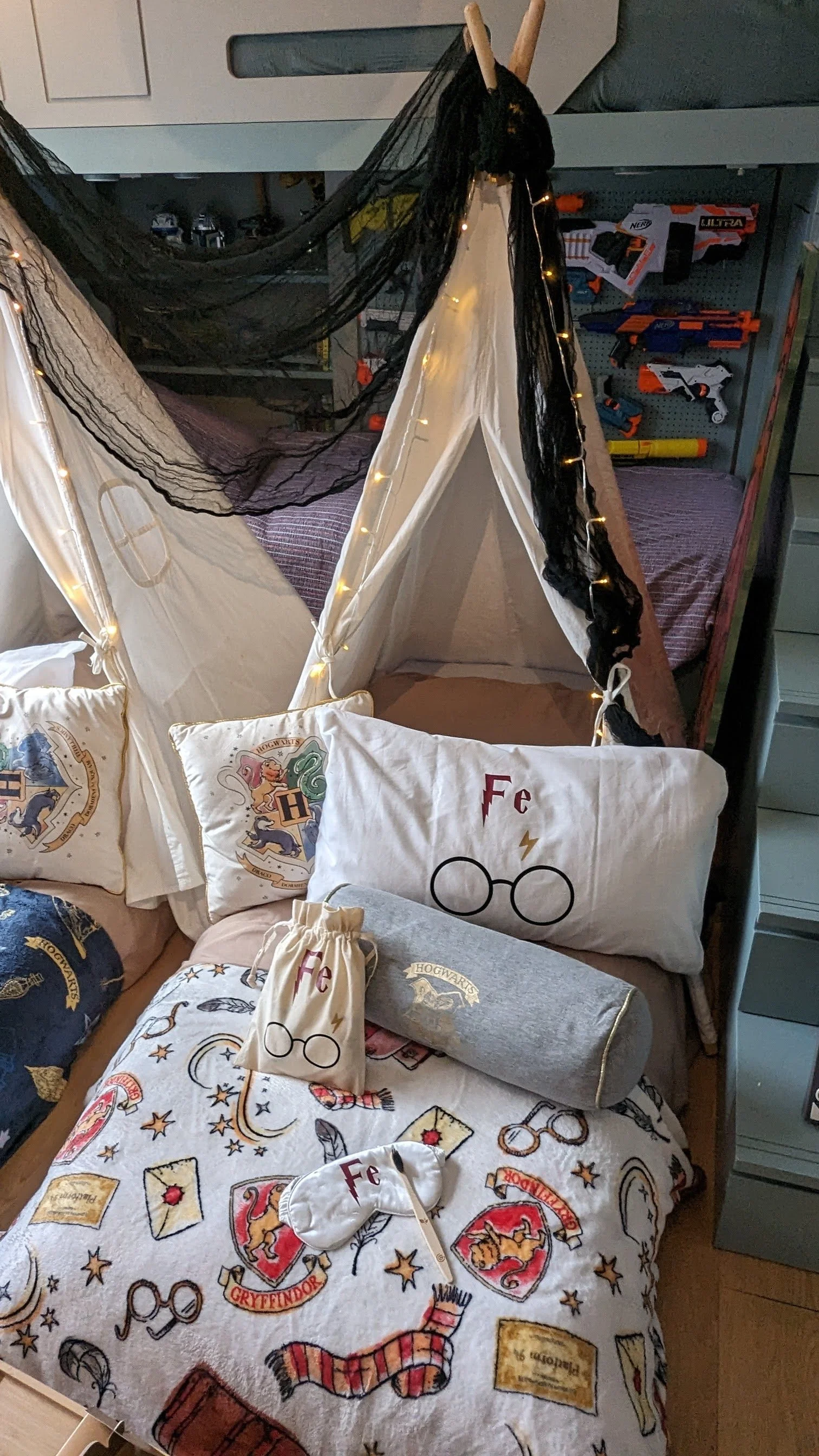 A children's bed decorated with Harry Potter and Hogwarts themed items, including pillows, a blanket, and a drawstring bag, with a canopy made of black and white fabric and string lights. A mattress with toys and a shelf of Nerf guns are visible in the background.