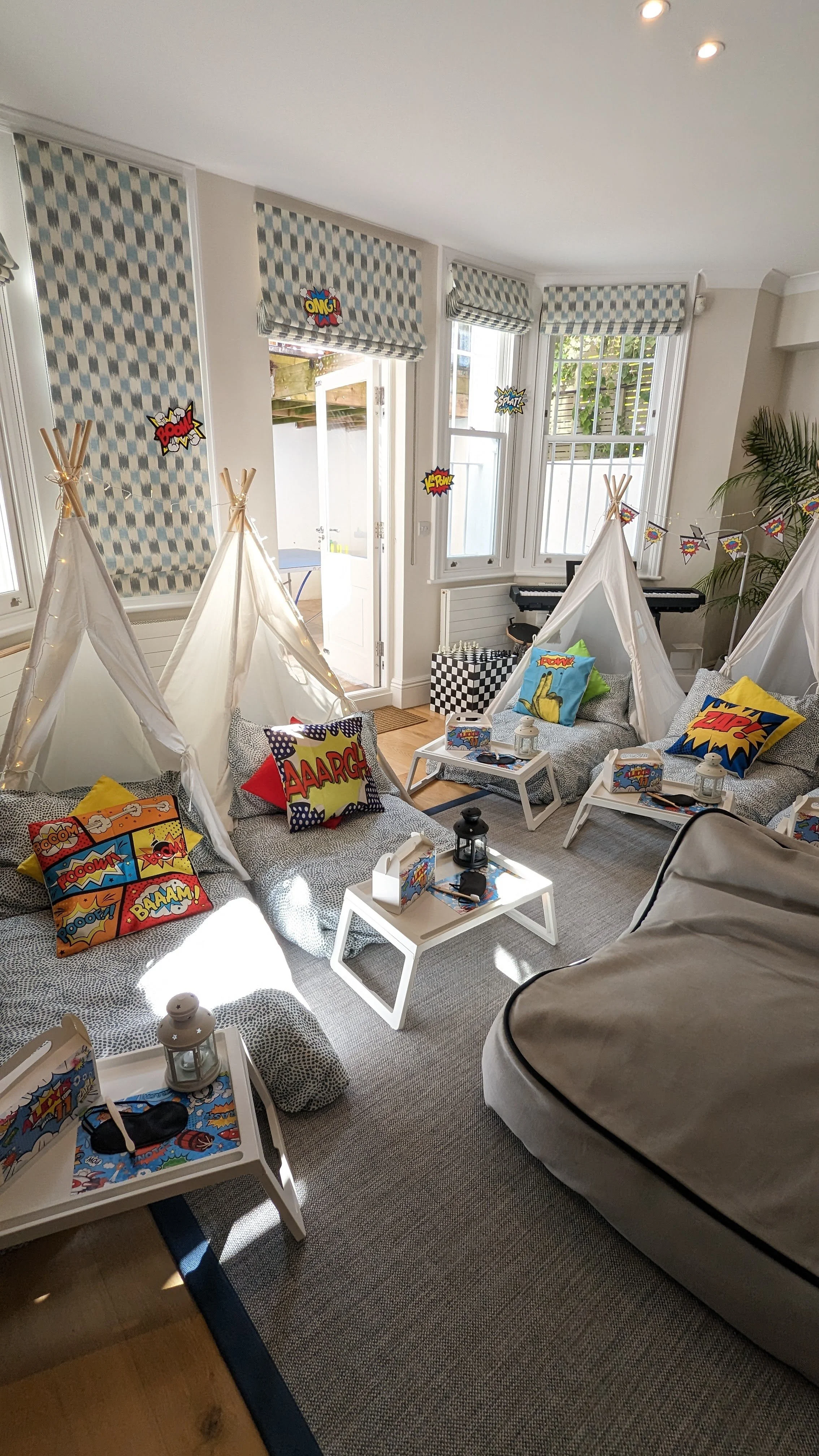 A playroom decorated with comic book style theme, featuring small teepee tents, superhero pillows, comic book wall art, and small tables with lanterns, in front of large windows with patterned window covers.