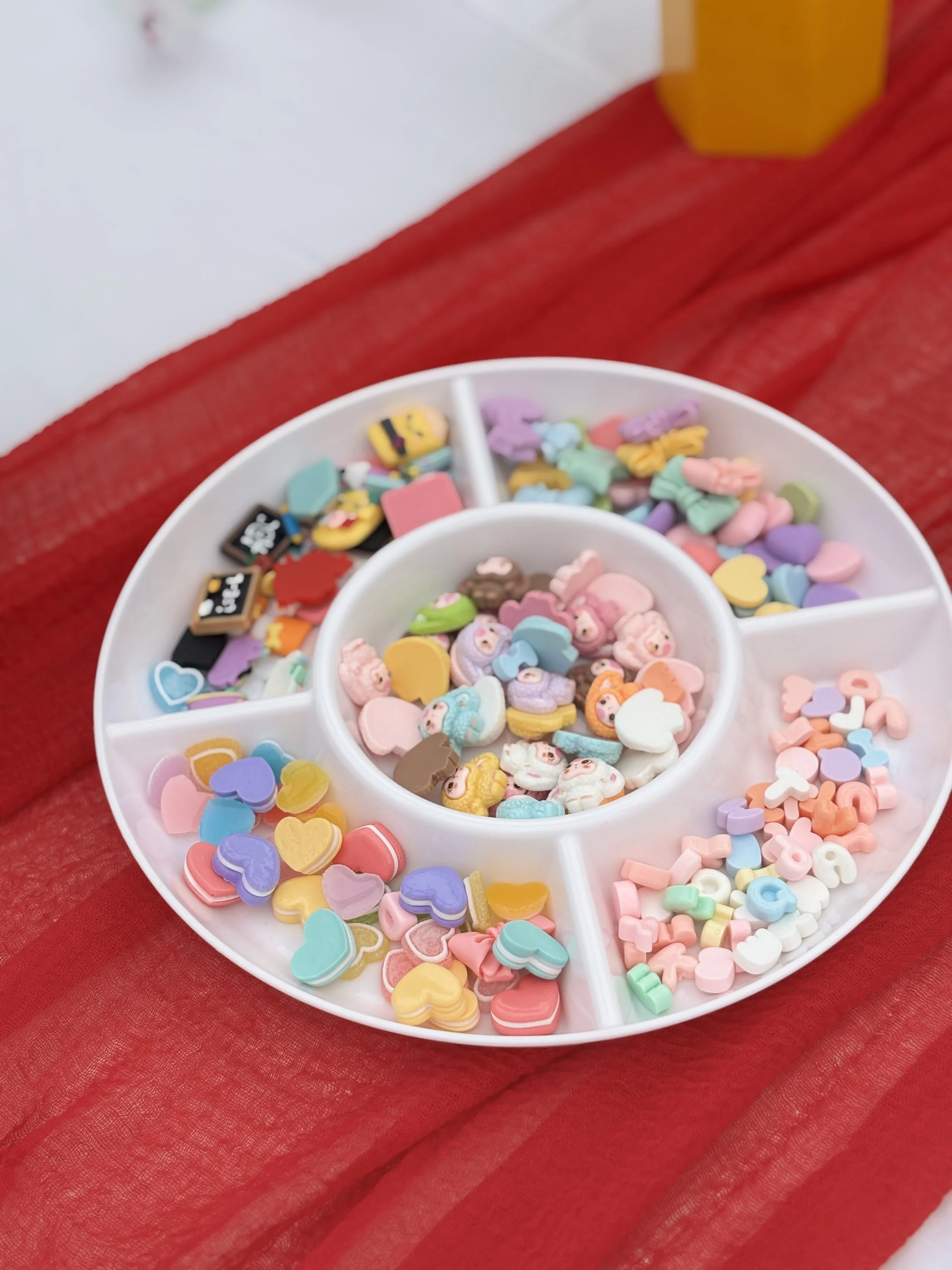 Colorful foam and rubber decorations in a white circular organizer with multiple compartments on a red fabric surface.