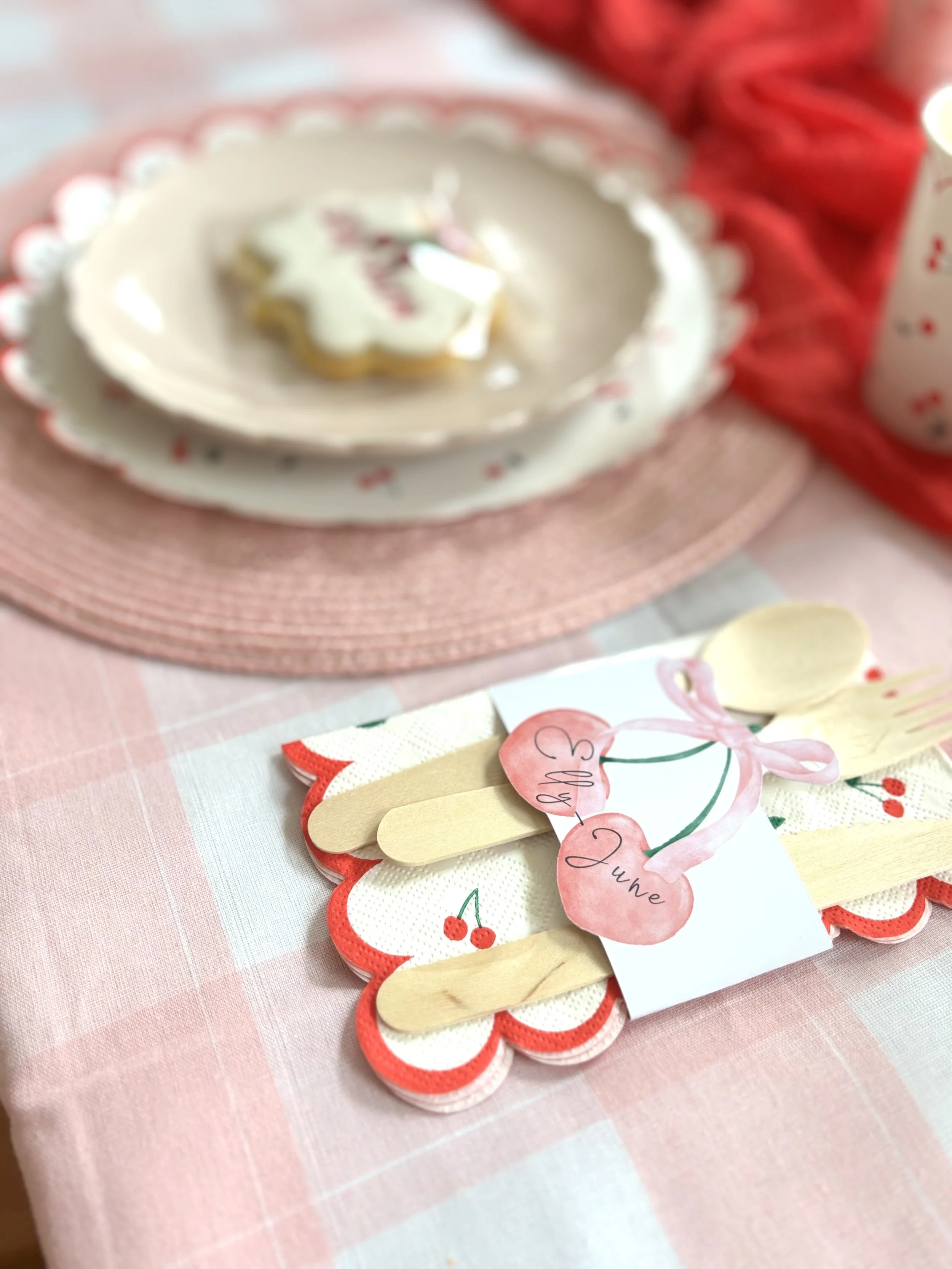 A Christmas-themed table setting with a pink checkered tablecloth, a pink placemat, a white plate with a star-shaped cookie decorated with white icing, and a paper cutout craft with cherries and pink bows.