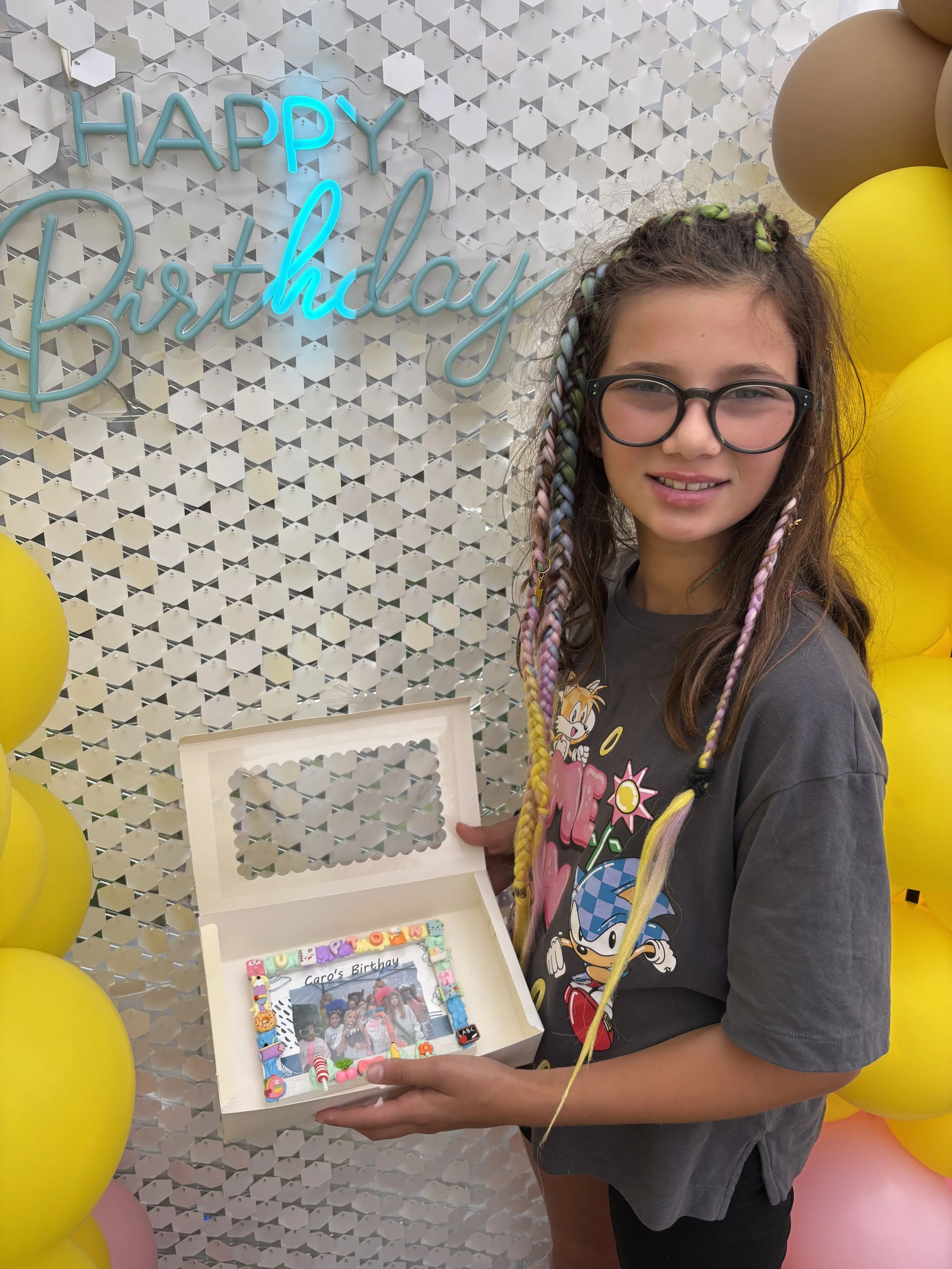 A girl with glasses holding a birthday card and decorations in front of a 'Happy Birthday' sign and yellow balloons.