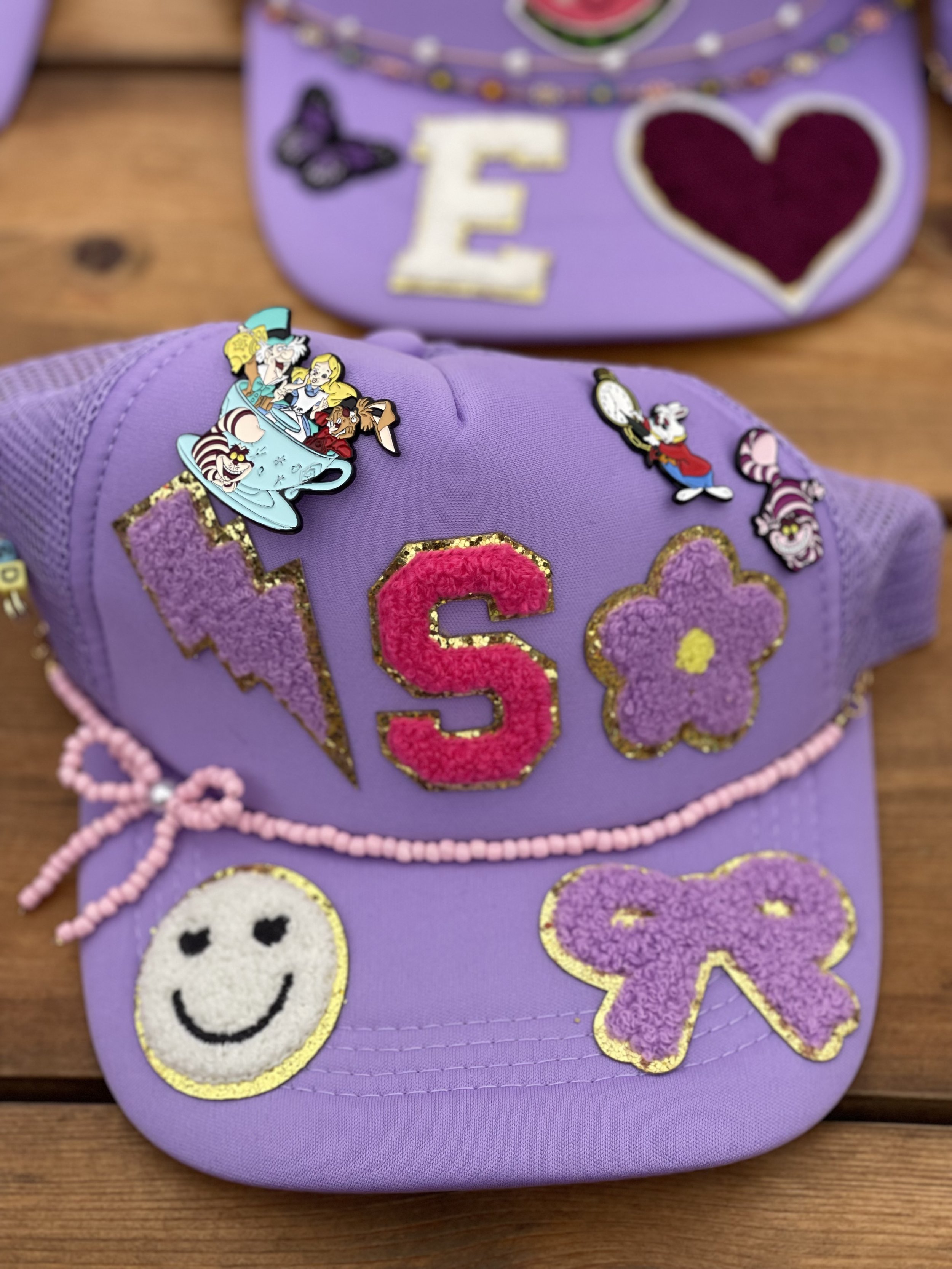 Purple cap decorated with glittery patches, including a lightning bolt, the letter 'S', a flower, a smiley face, and a bow. Also features various Disney character pins.