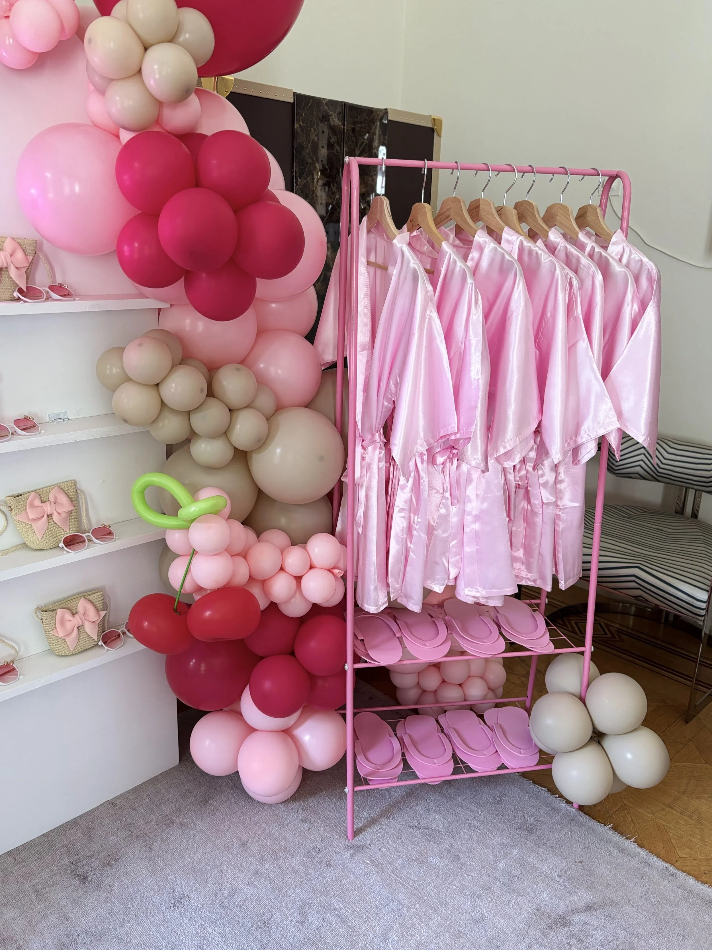 Pink satin bathrobes hanging on a pink clothing rack, pink slippers on the shelves below, surrounded by pink, beige, and white balloons, with small beige bags with pink bows on the shelves to the left.